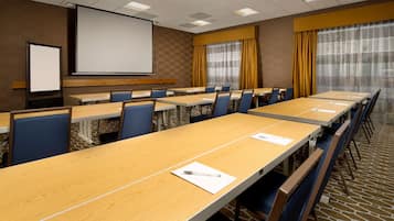 A conference room with long tables, chairs, a projector screen, and a large window with curtains.