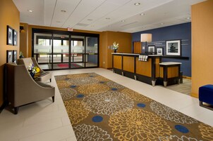 Un vestíbulo de hotel con mostrador de recepción, una sala de espera con sillas, alfombra estampada y cuadros en las paredes.