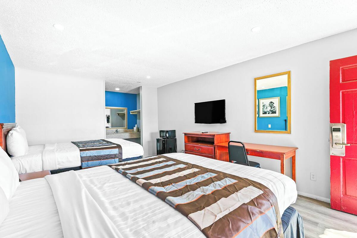 A hotel room with two beds, a desk, a chair, a TV, and a red door.