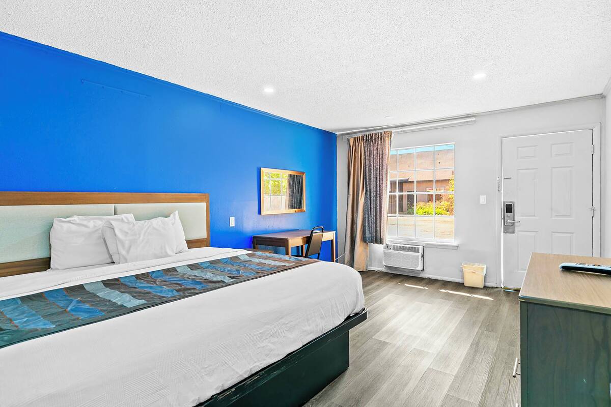 A hotel room with a large bed, a desk, a chair, a window with curtains, and a door.