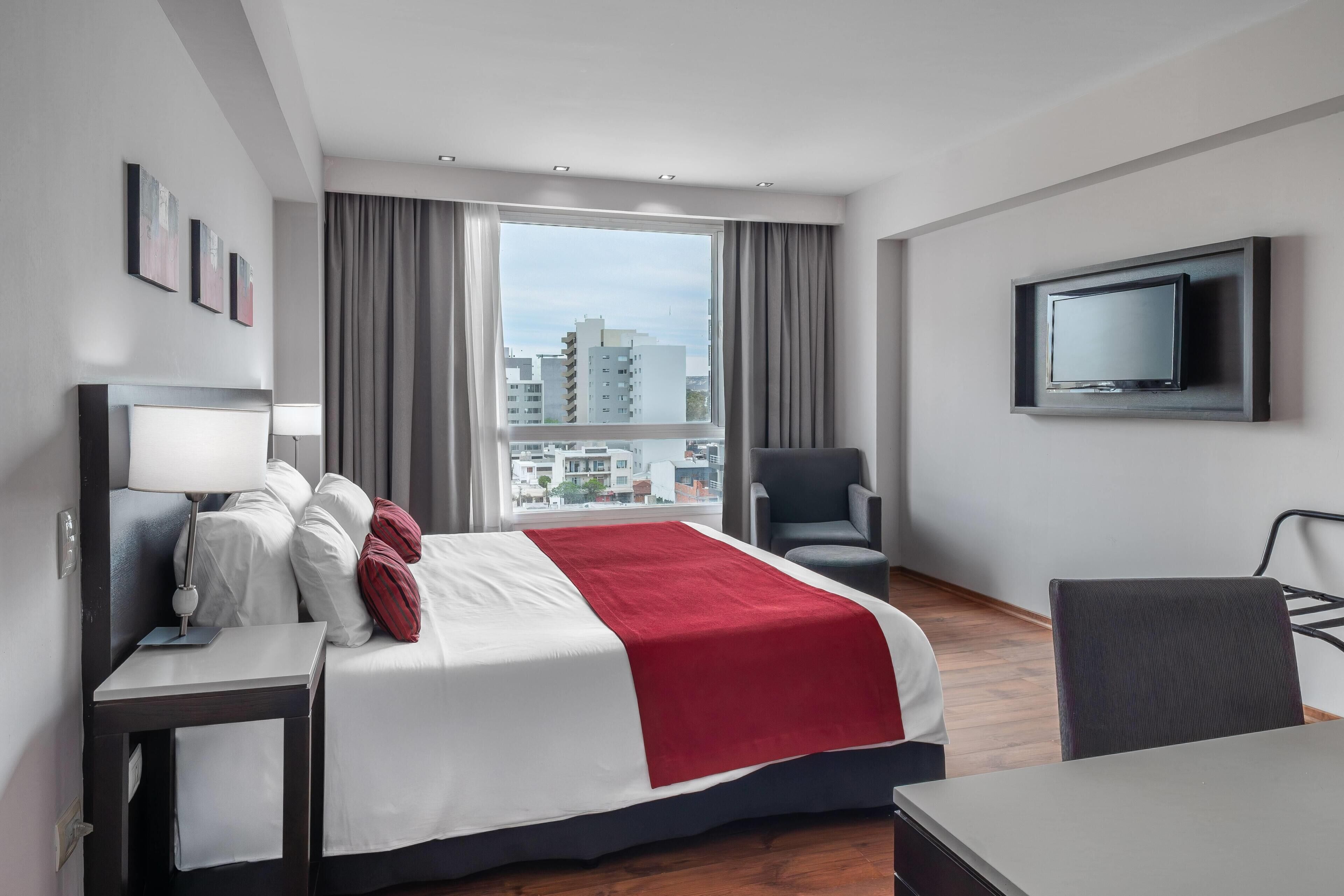 Habitación de hotel con cama, escritorio, silla, televisor y una ventana grande con vistas a la ciudad.