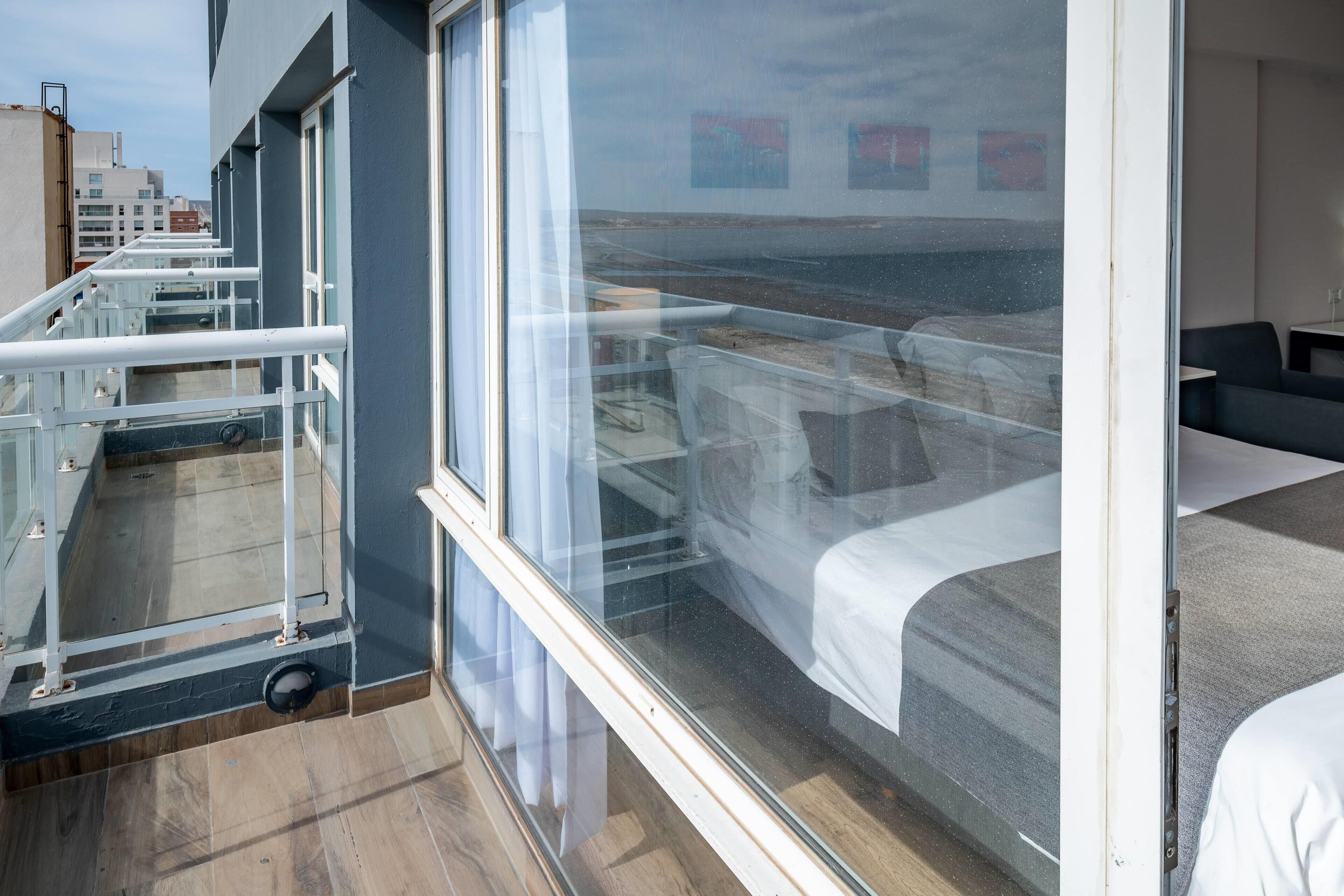 Un balcón con barandilla de vidrio, una cama con ropa blanca y vistas de la ciudad.