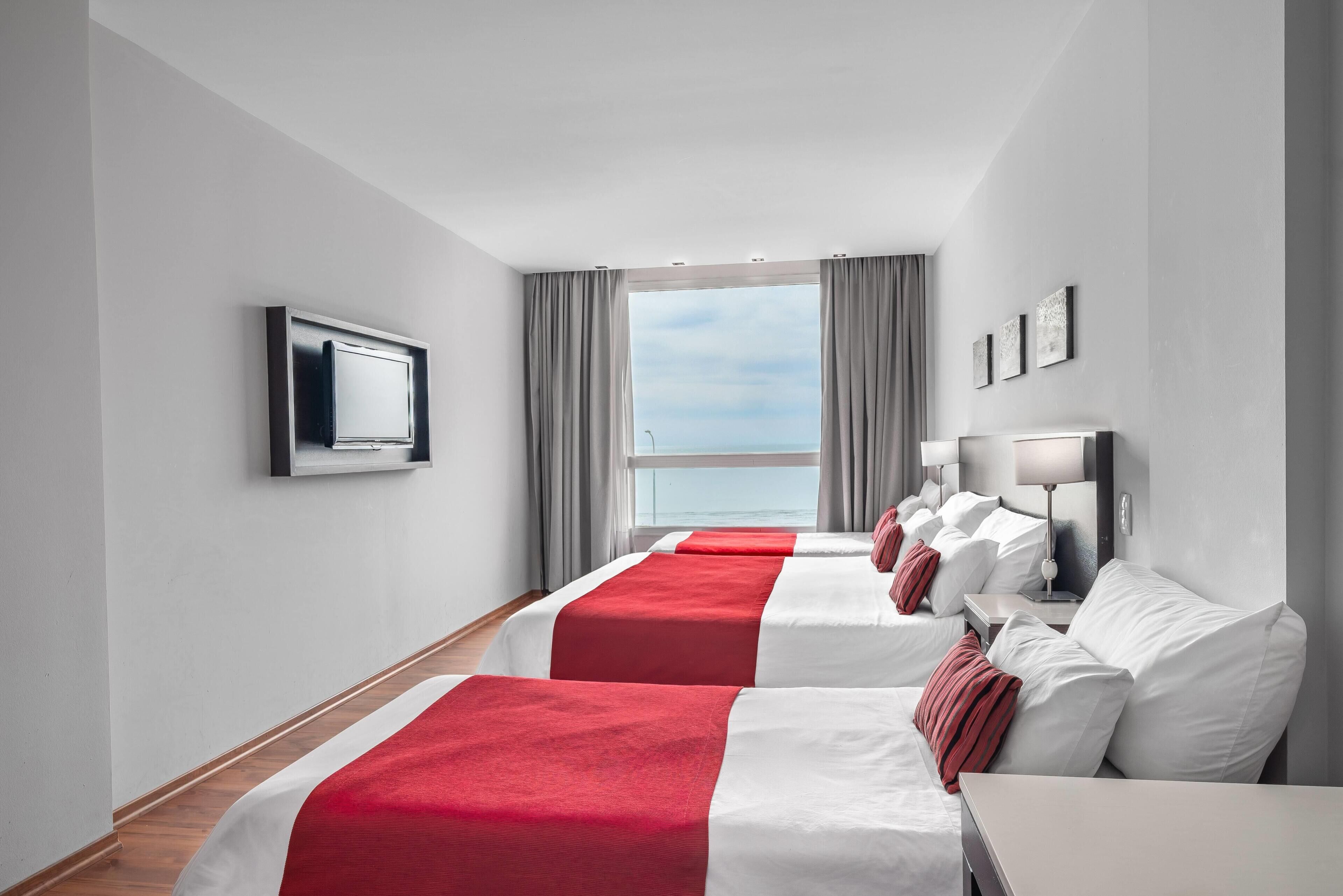 Quarto de hotel com duas camas, televisão, janela com cortinas e vista para o mar.
