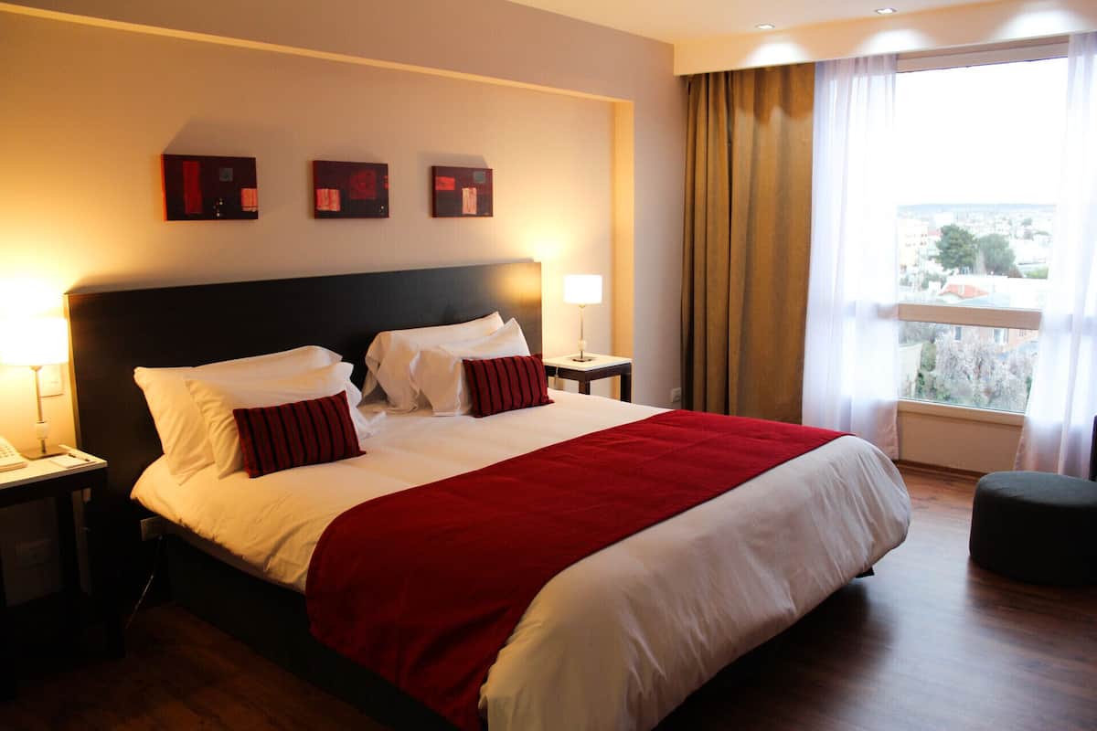 A hotel room with a large bed, two bedside tables, a window with curtains, and artwork on the wall.