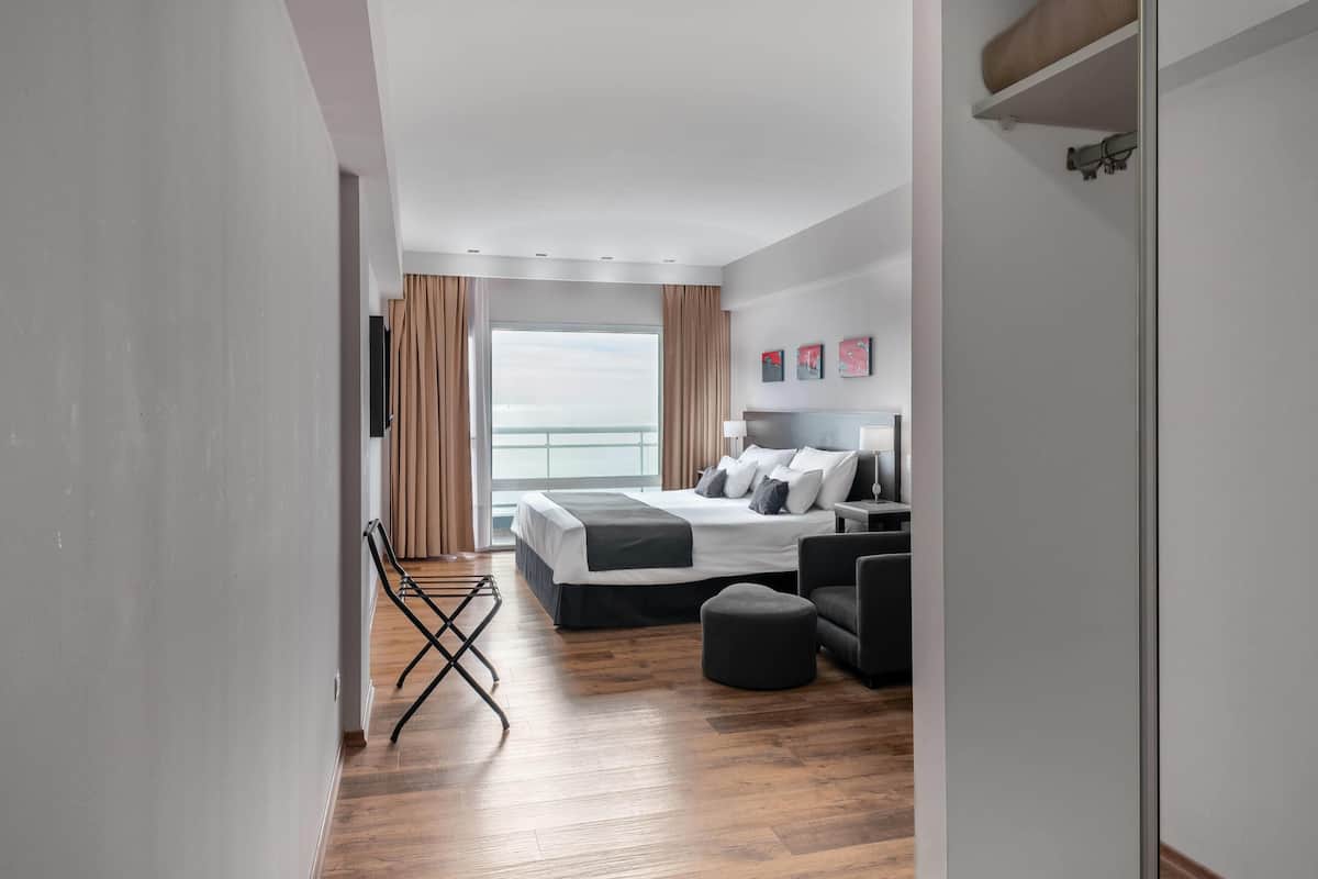 A modern hotel room with a large bed, a sofa, a small table, and a view of the sea.