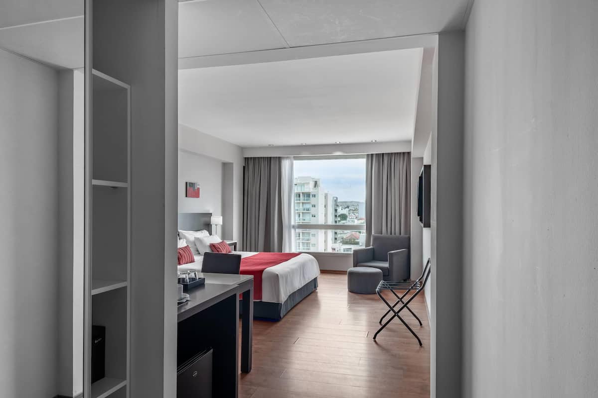 A modern hotel room with a bed, a desk, a chair, and a view of the city.