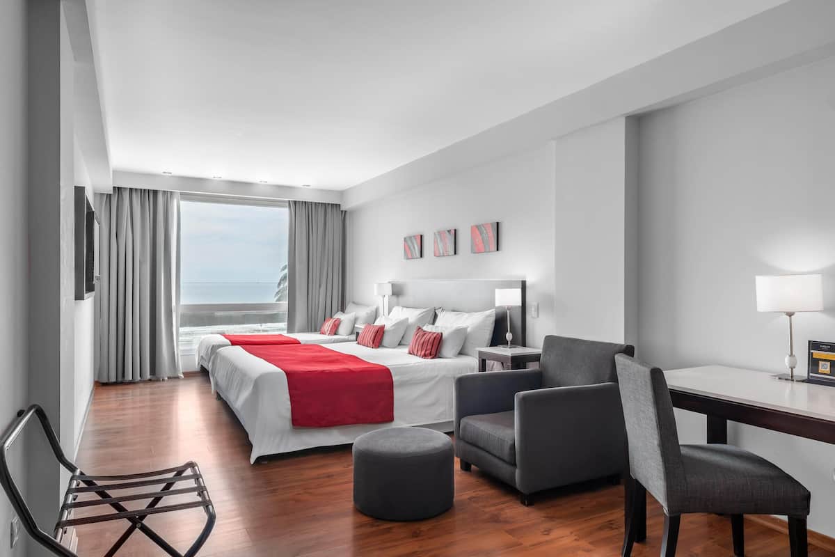 A hotel room with a bed, two chairs, a desk, a lamp, and a view of the sea.