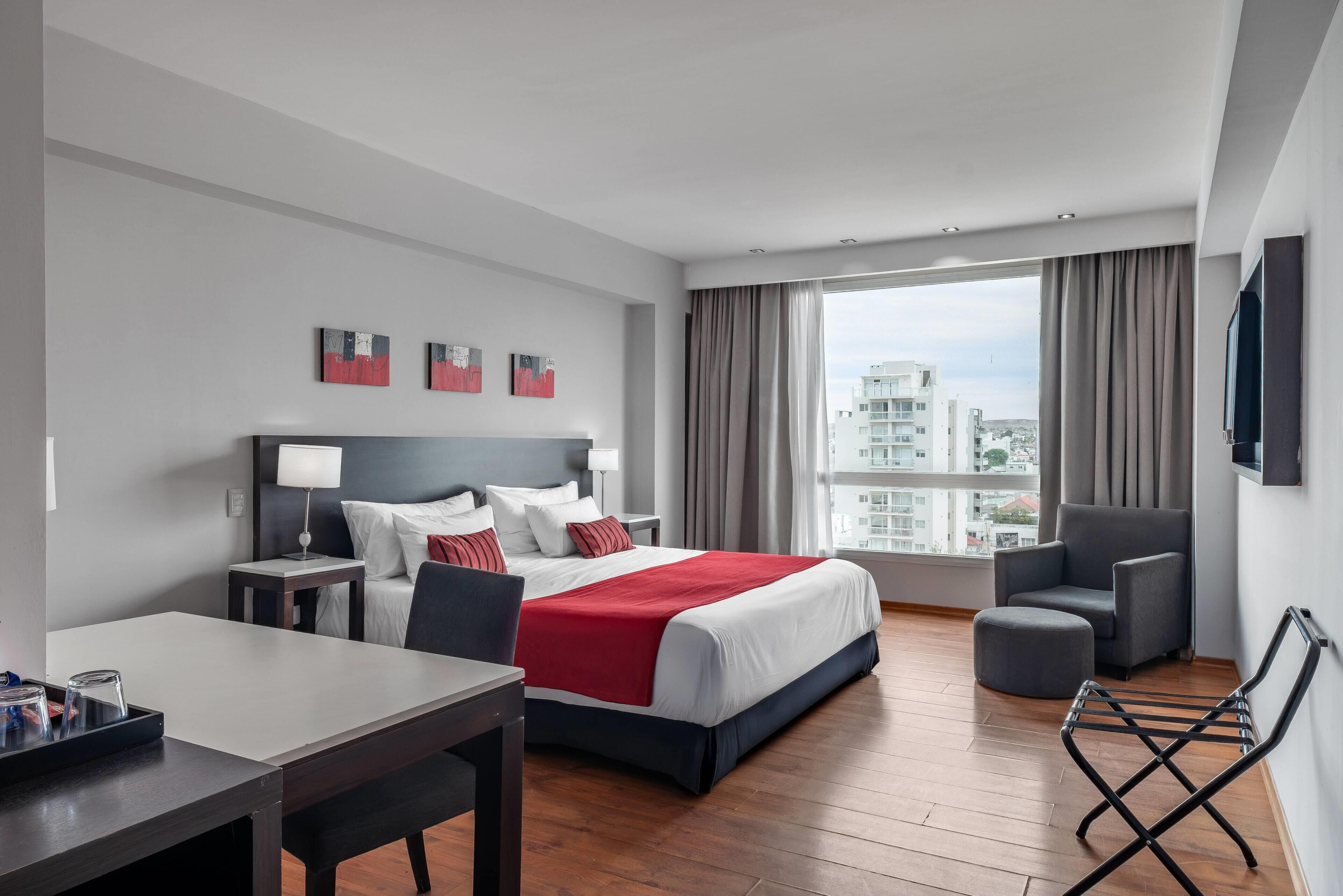 A modern hotel room with a large bed, a desk, a chair, and a view of the city.