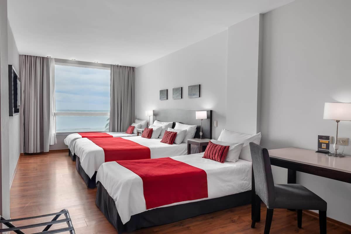A hotel room with two beds, a desk, a chair, and a view of the sea.