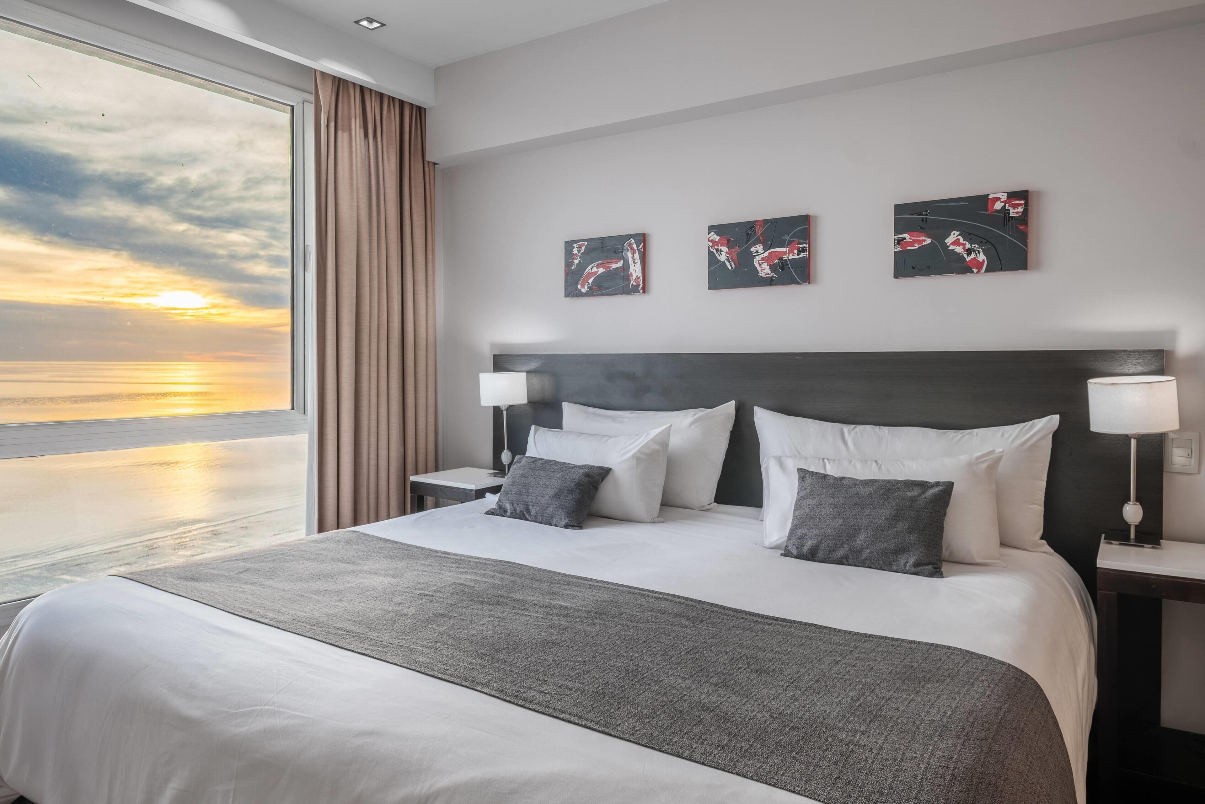 Hotelkamer met een groot bed, twee nachtkastjes, uitzicht op de zonsondergang en drie kunstwerken aan de muur.