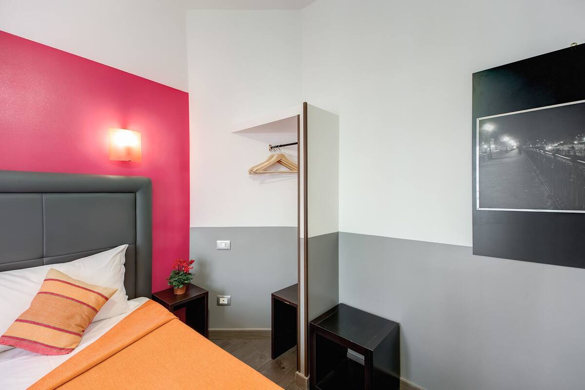 Una habitación de hotel moderna con cabecera gris, cama con ropa de cama naranja, pared de acento rosa, luz empotrada, un perchero de madera, una mesita pequeña y un marco de fotos negro.
