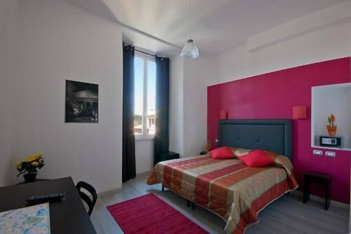 A bedroom with a bed, a desk, a chair, a window, and a wall clock.