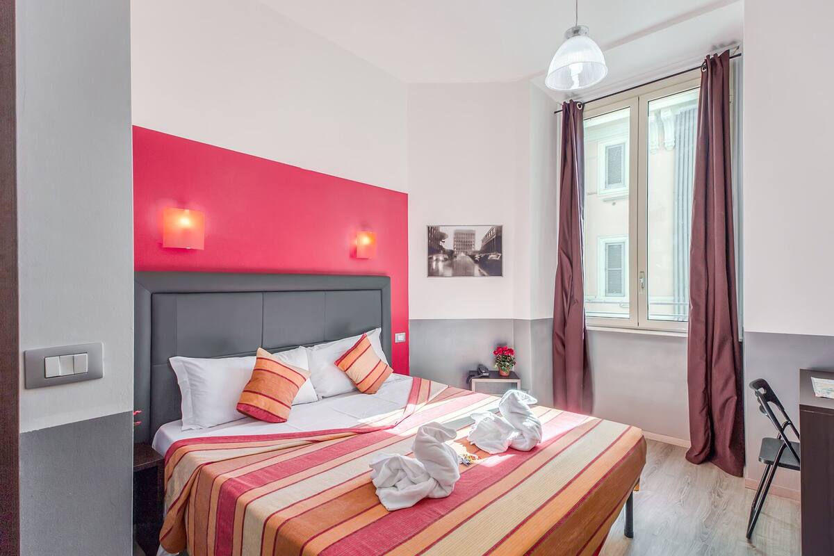 Una habitación de hotel con cama, mesita de noche, lámpara y ventana con cortinas.