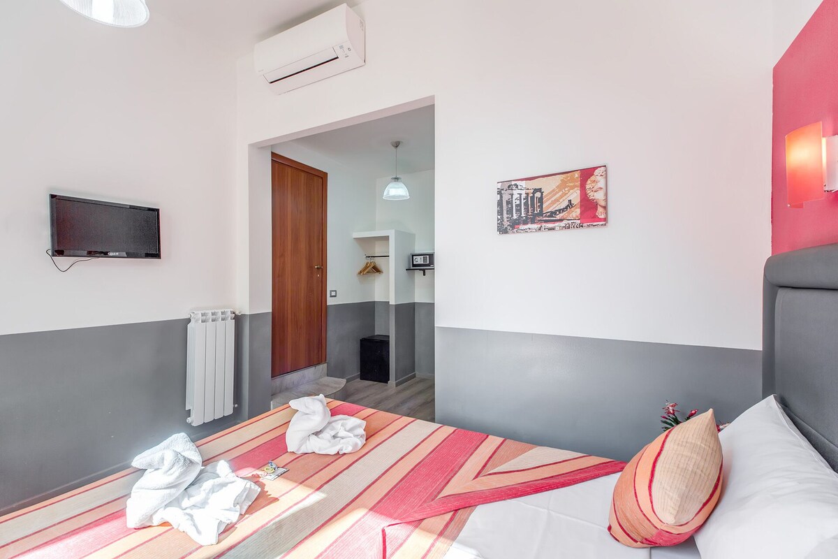 A hotel room with a bed, a flat-screen TV mounted on the wall, a radiator, and a door leading to a bathroom.