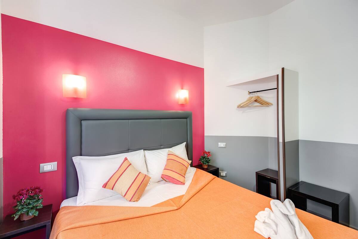 Una habitación de hotel con cama, mesita de noche, espejo y pared con una decoración en tonos rojo y gris.