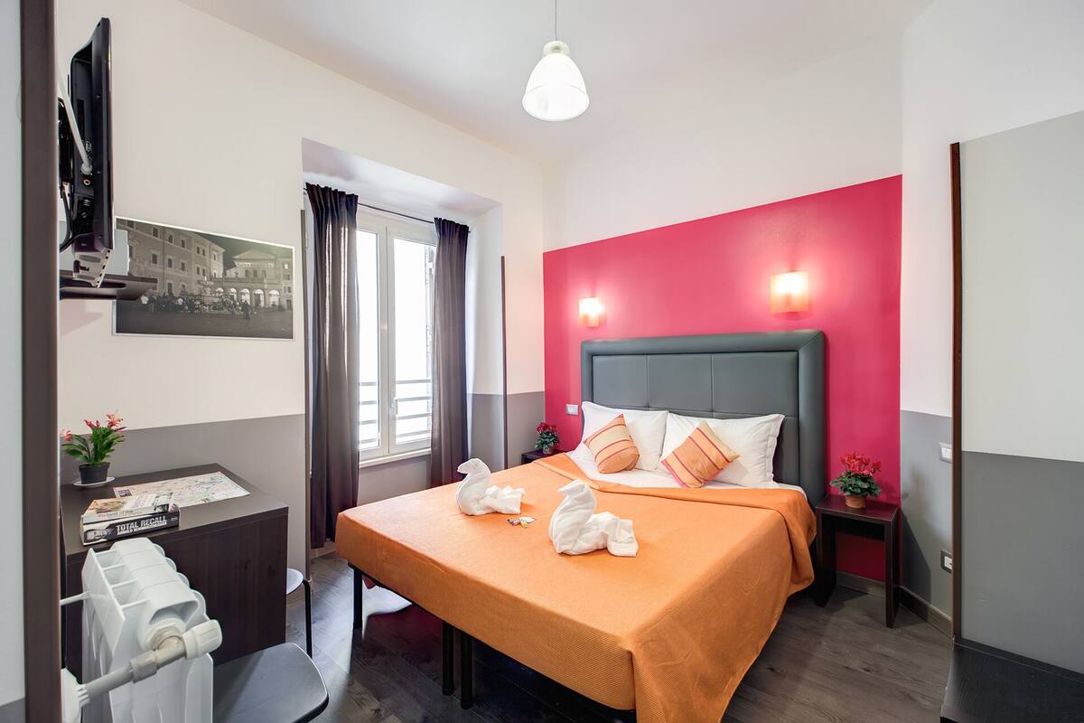 Habitación de hotel con cama, escritorio, silla, ventana con cortinas e imagen colgada en la pared.