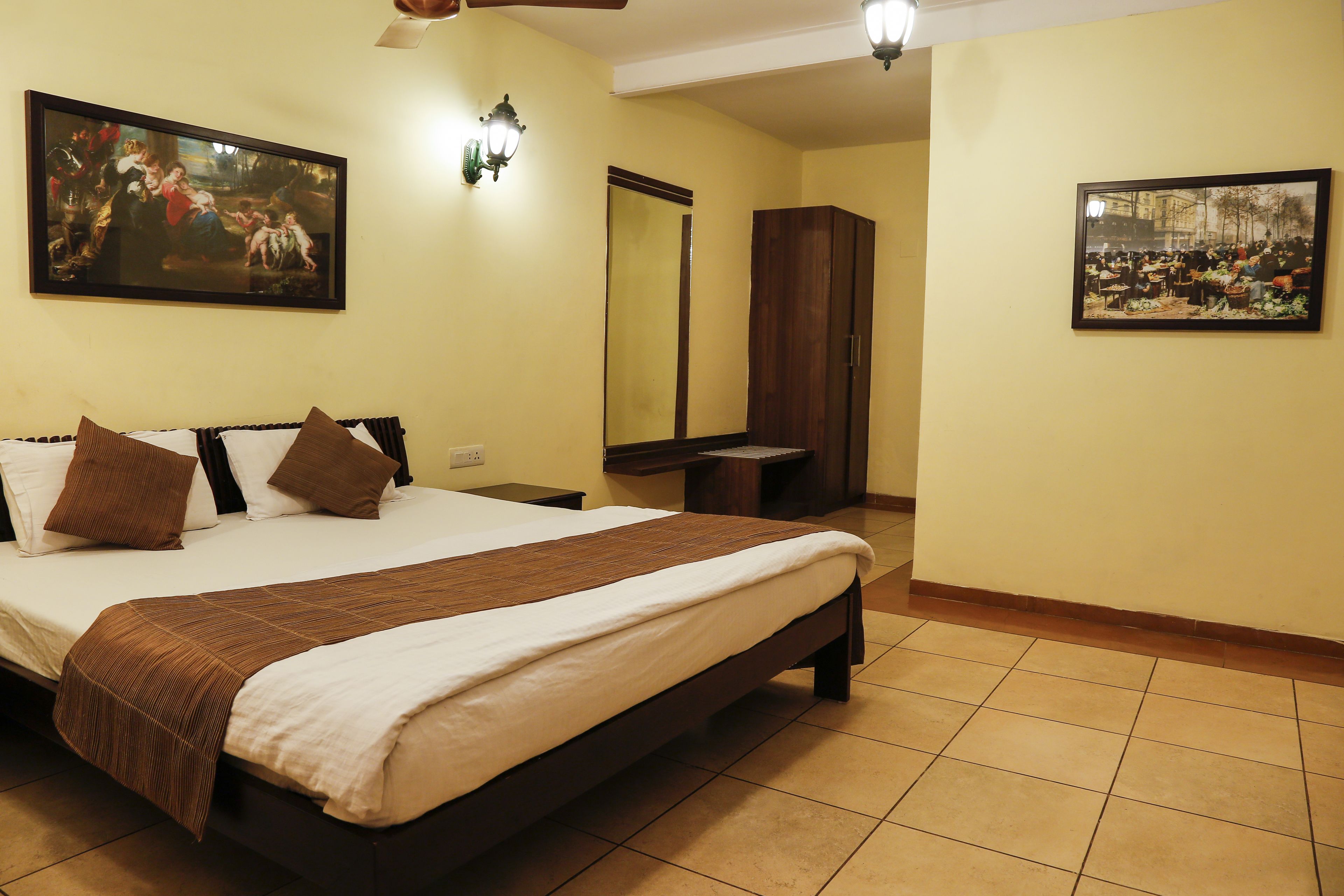A neatly made bed with a brown bedspread, two brown pillows, and a wooden headboard. There is a large framed painting on the wall above the bed and a mirror on the opposite wall.