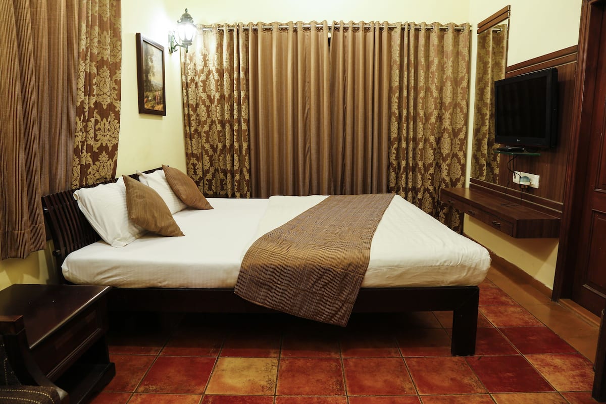 A neatly made bed with a brown blanket, a television mounted on the wall, and curtains with a floral pattern.