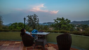 Une terrasse dotée de chaises et d’une table en osier, offrant une vue panoramique sur un paysage verdoyant, avec des arbres et des collines à l’horizon.