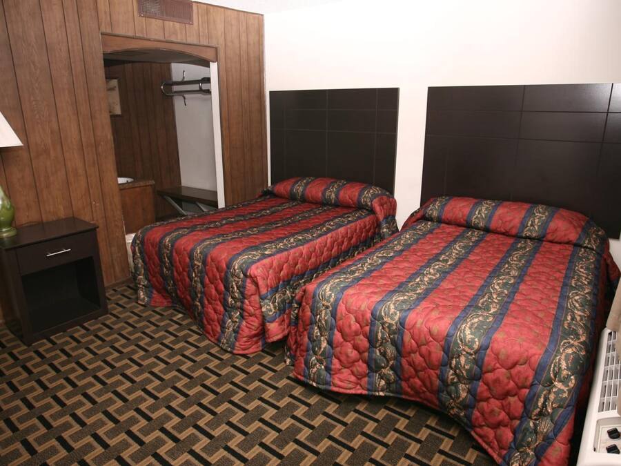Habitación de hotel con dos camas, un armario de madera, una mesita de noche con lámpara y suelo estampado.