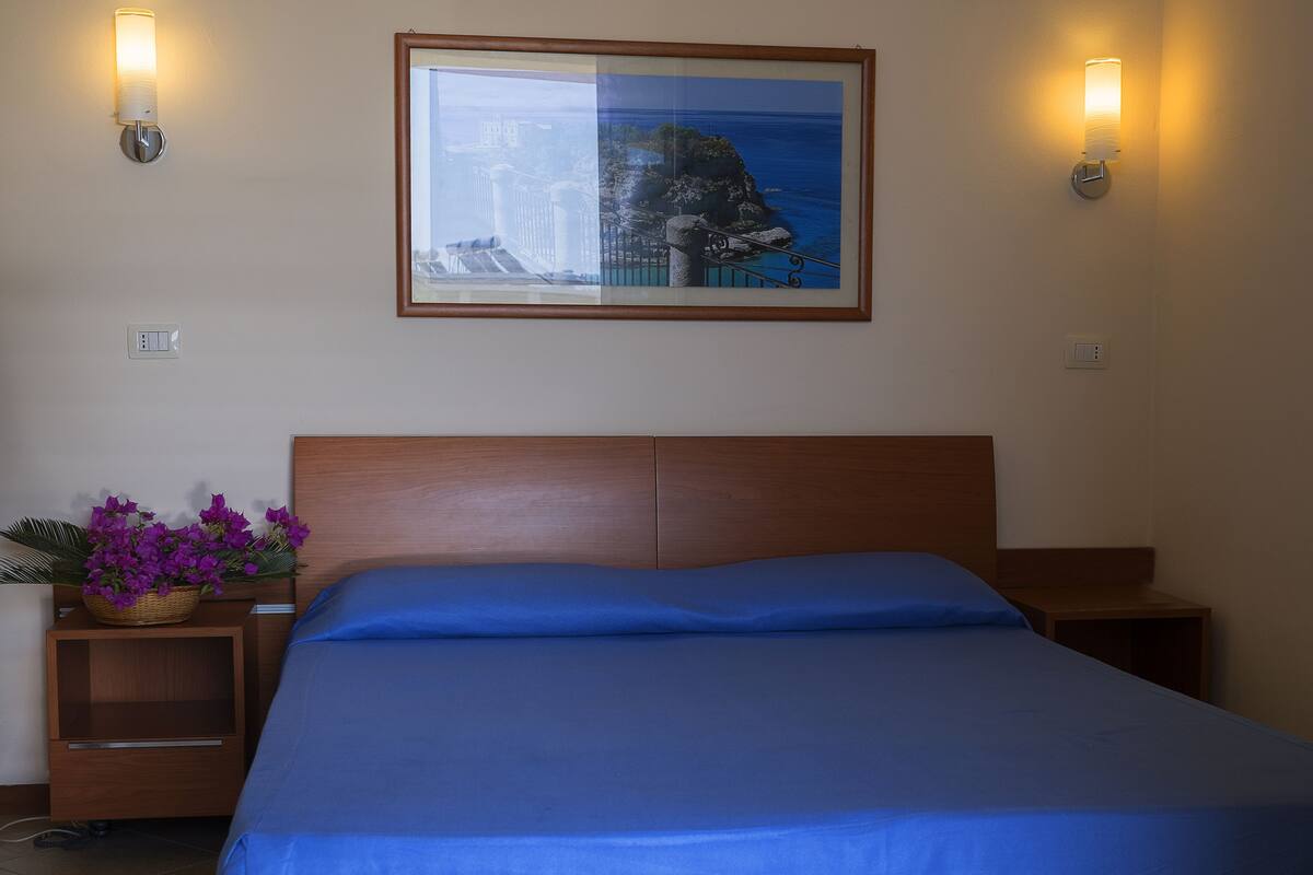 A hotel room with a bed, a nightstand with a lamp, a framed picture on the wall, and a basket of flowers.