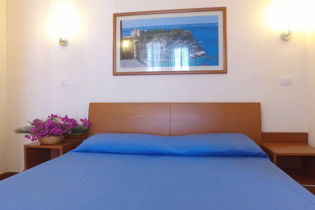A bedroom with a bed, a framed picture of a coastal scene, and a nightstand with purple flowers.