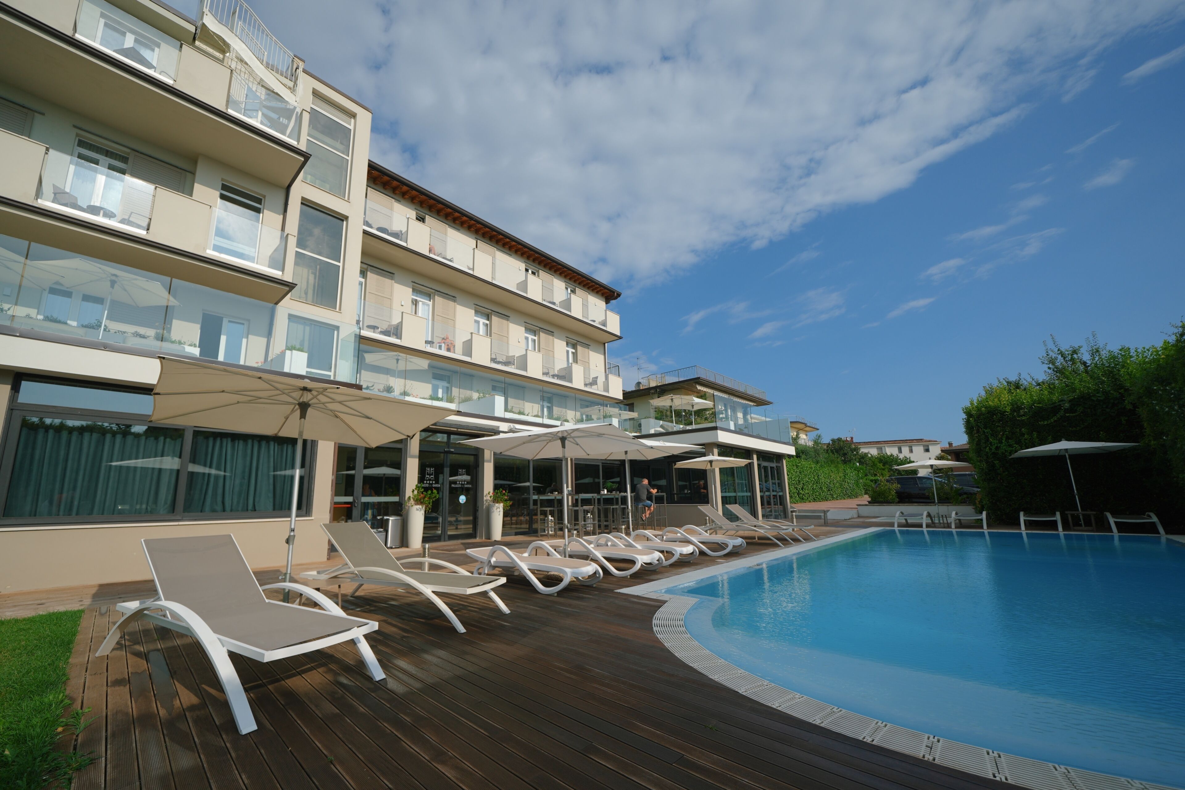 Un área junto a la piscina con sillones de descanso y sombrillas, una terraza de madera y un edificio con balcones.