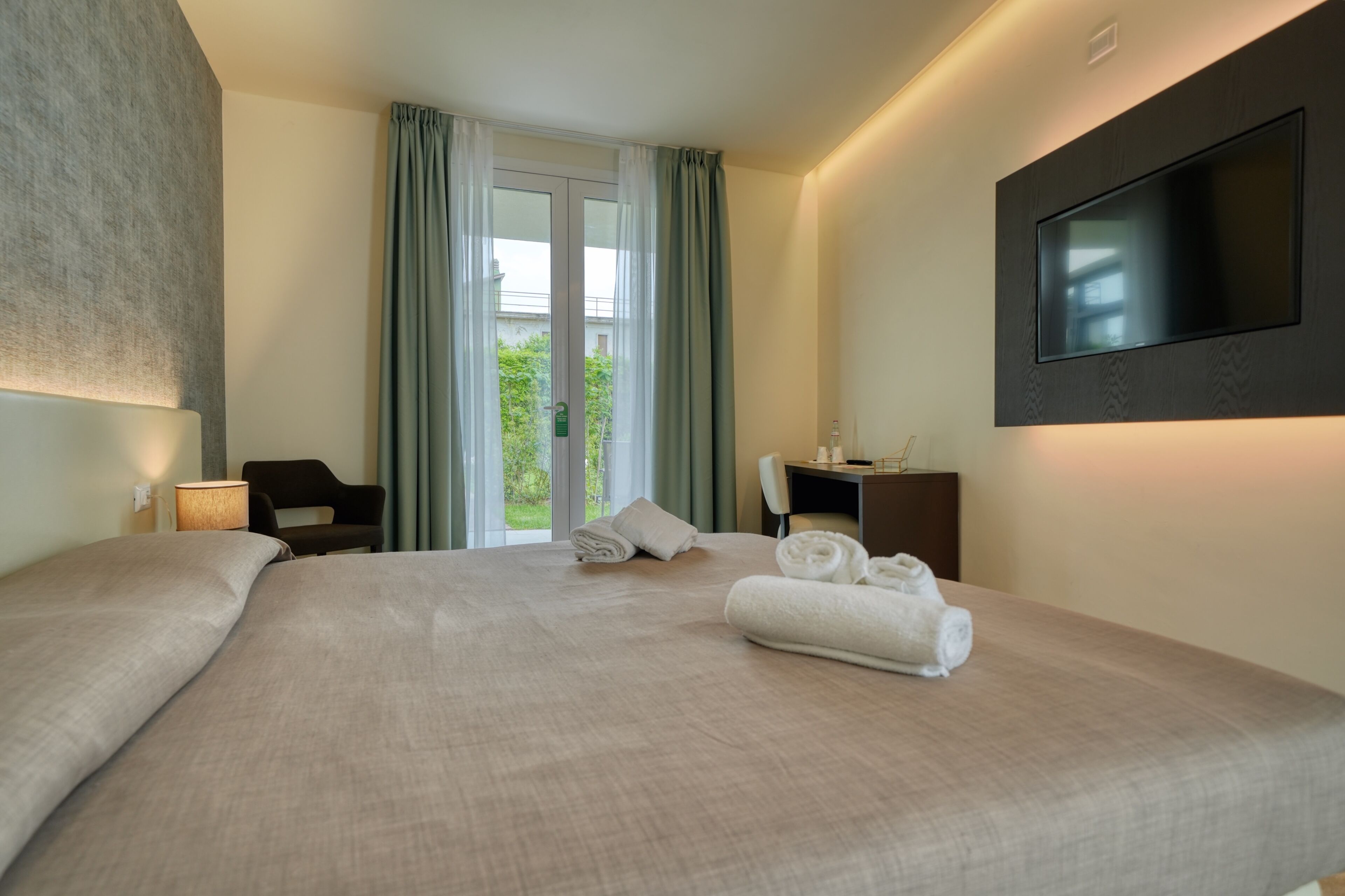 A hotel room with a large bed, a desk, a chair, a TV mounted on the wall, and a view of greenery outside.