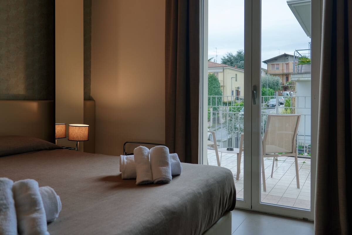 Una habitación de hotel con cama, una mesita de noche con lámpara y vistas al balcón exterior.