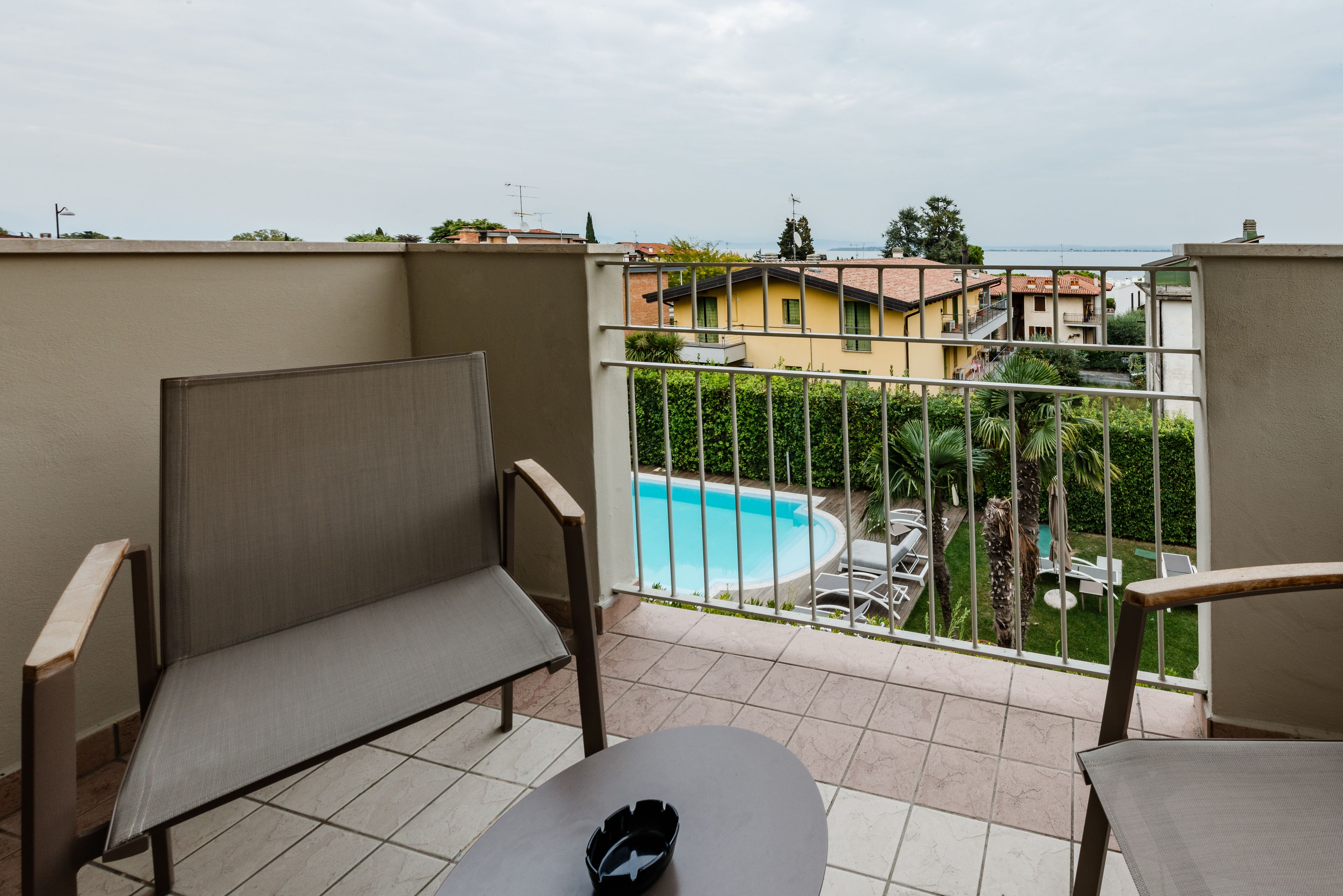 Ein Balkon mit zwei Stühlen, einem Tisch und Blick auf ein Schwimmbecken sowie Gebäude im Hintergrund.
