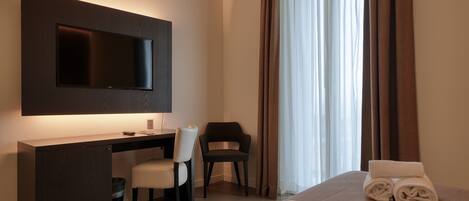 Ein Hotelzimmer mit Flachbildfernseher an der Wand, einem Schreibtisch mit Stuhl, einem Bett mit zusammengerollten Handtüchern und einem großen Fenster mit Vorhängen.