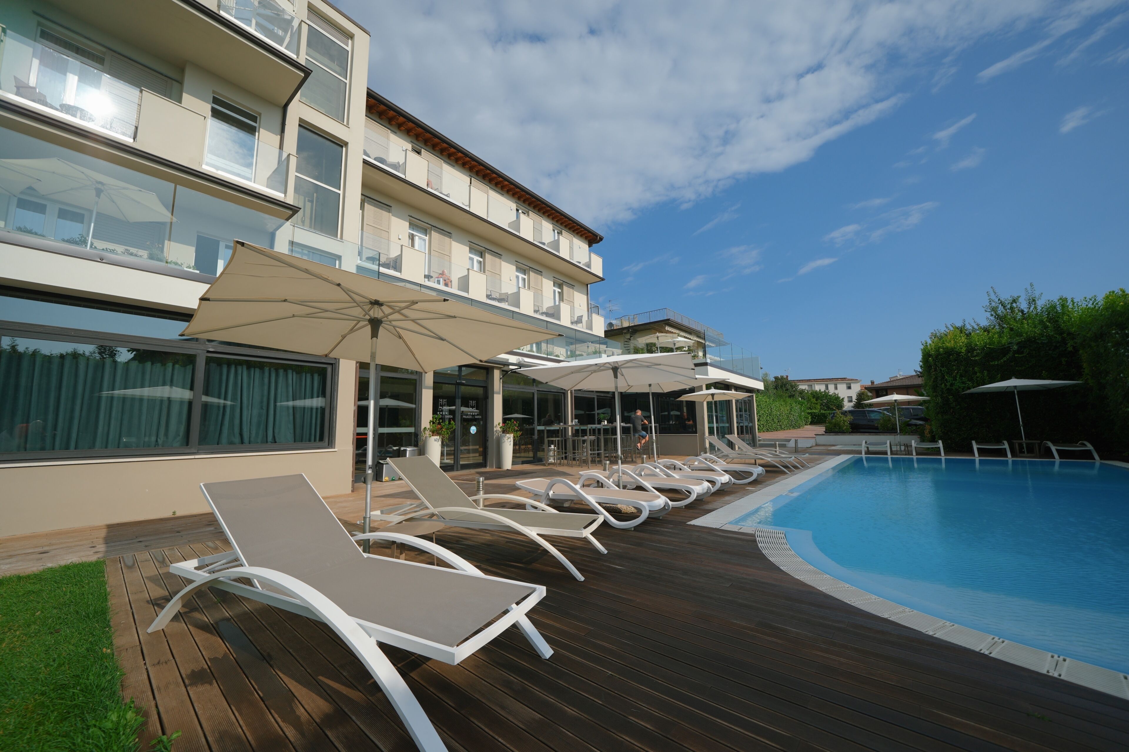 A poolside area with lounge chairs and umbrellas, a wooden deck, and a building with balconies.