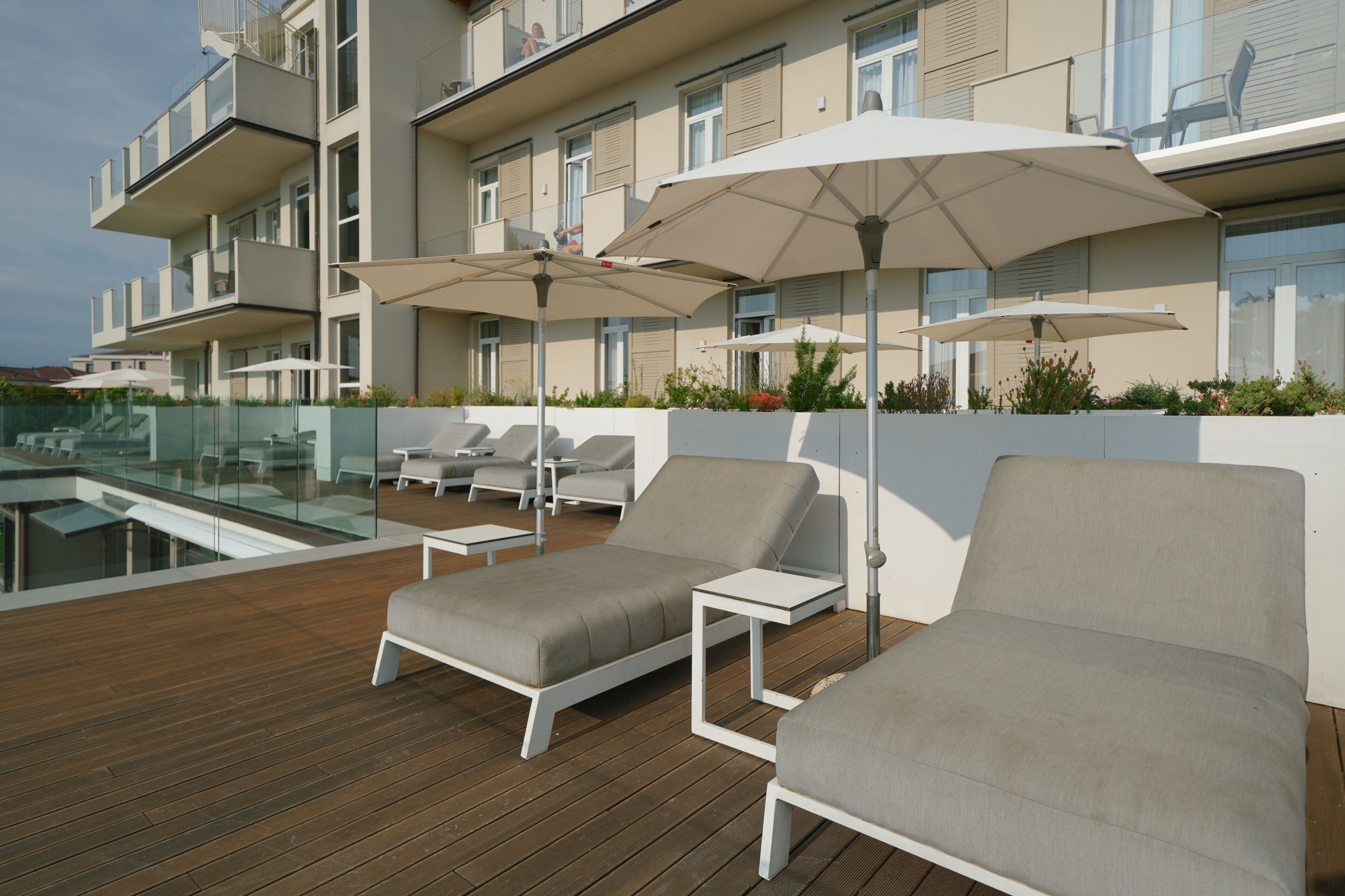 Un área de lounge en la azotea con tumbonas y sombrillas, una piscina y un edificio moderno con balcones.