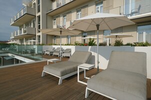 Un área de lounge en la azotea con tumbonas y sombrillas, una piscina y un edificio moderno con balcones.