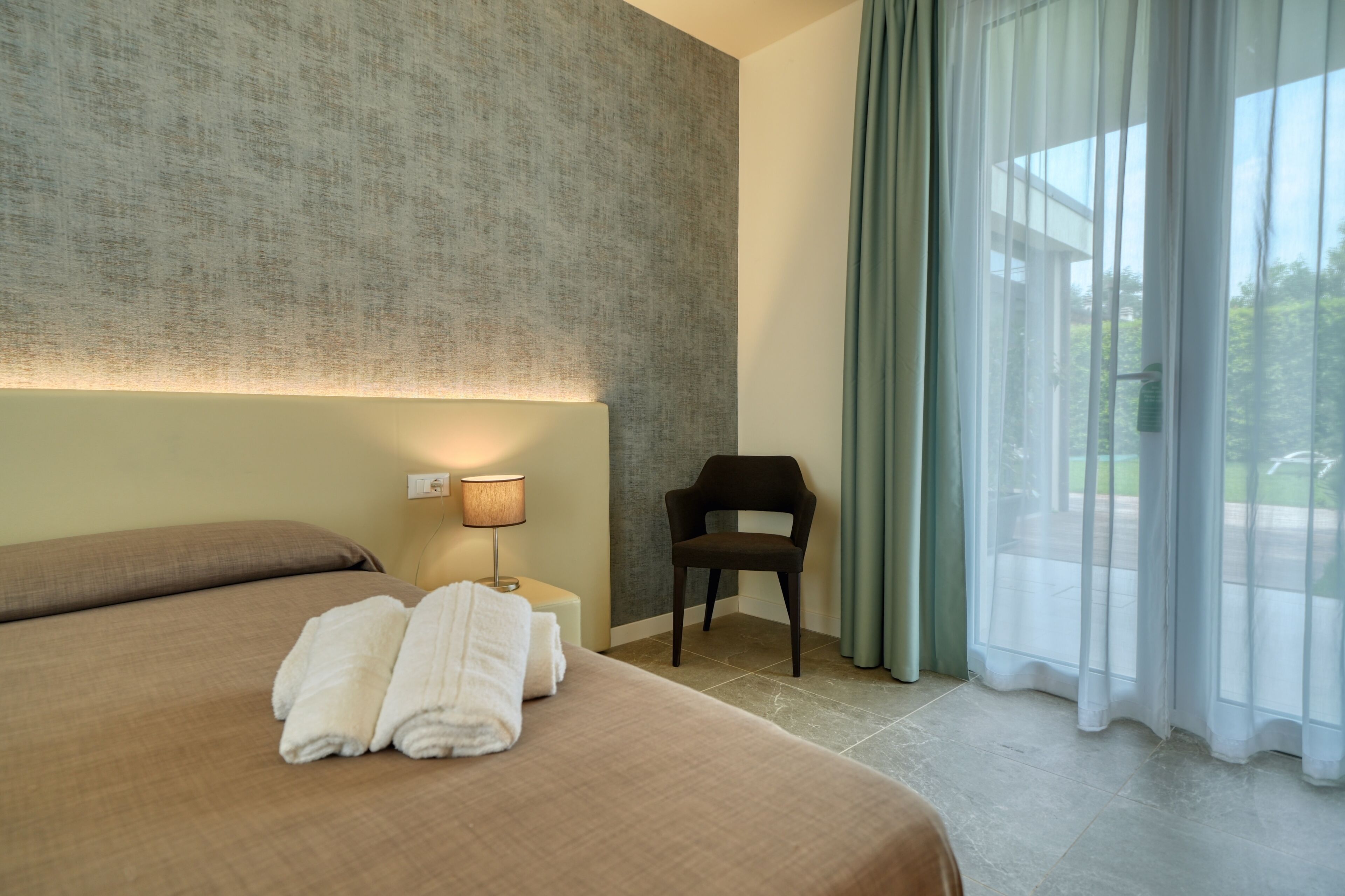 A hotel room with a large bed, a chair, a lamp, and a view of the outdoors.