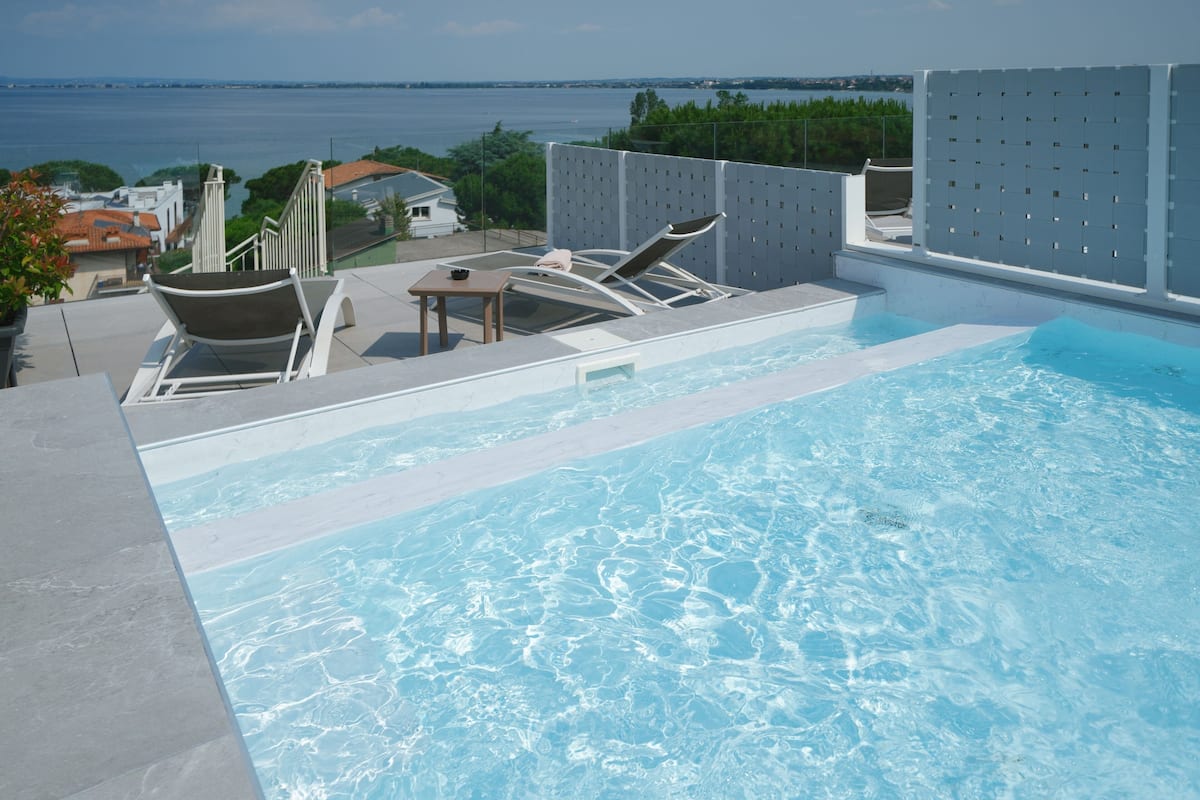 Una piscina infinita en la azotea con sillones de descanso y una mesa, con vistas a una masa de agua y vegetación.