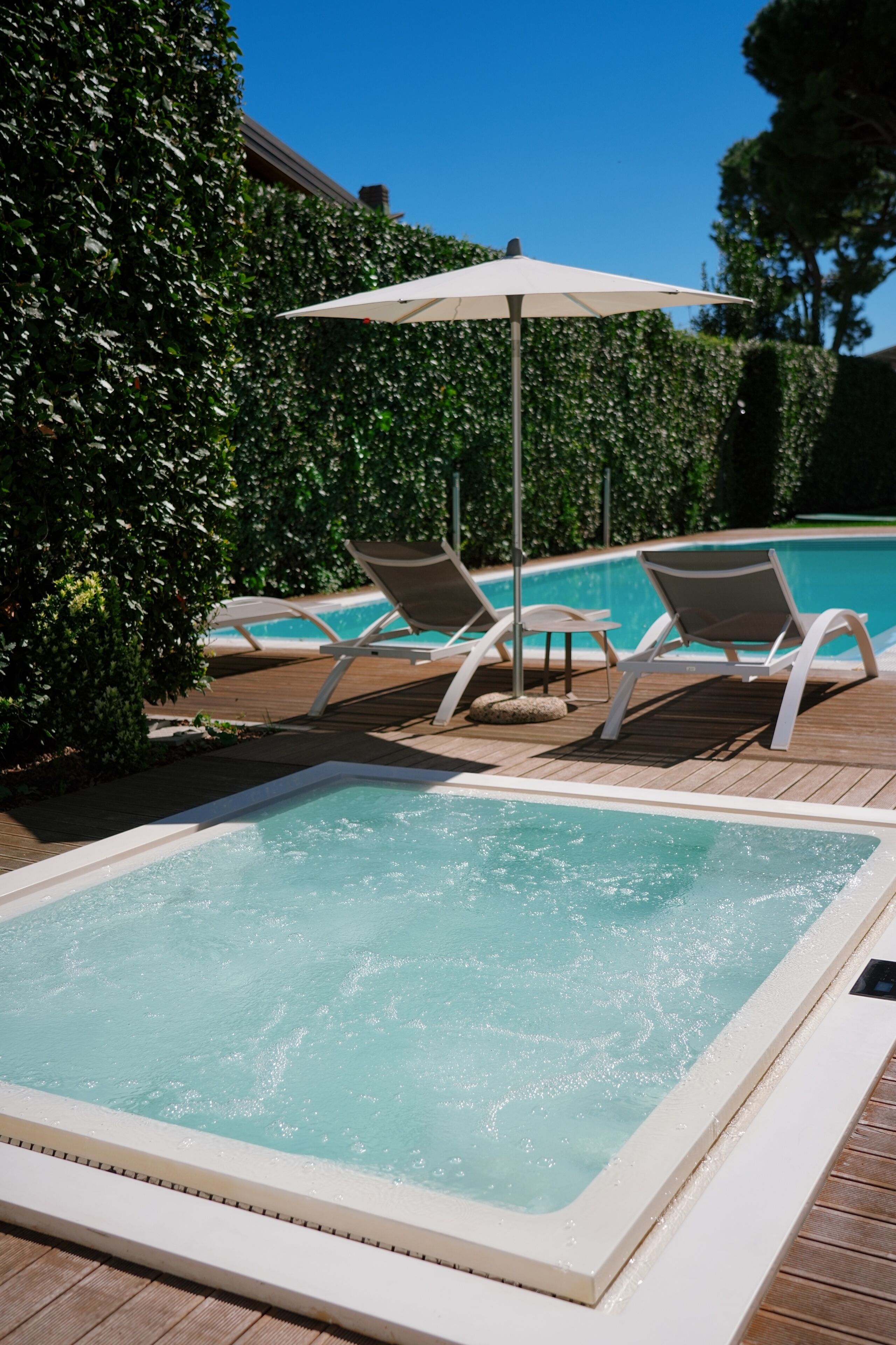 Een hottub met lounge fauteuils en een parasol, omringd door een houten terras en groen.