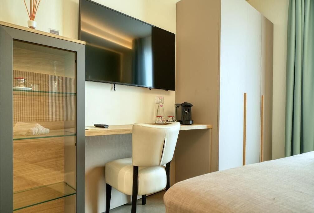 Ett hotellrum med en säng, ett skrivbord med en stol, en tv som är monterad på väggen, en kaffemaskin och ett glasskåp.