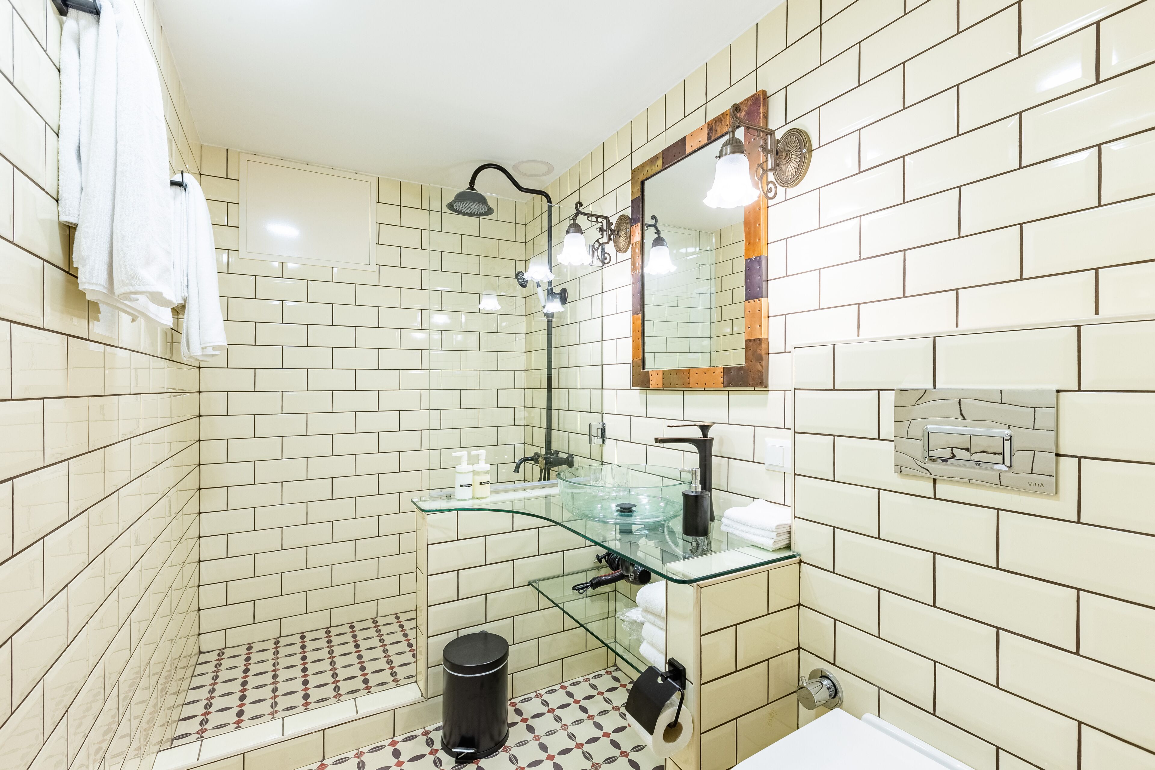 A modern bathroom with white subway tiles, a glass shower enclosure, a glass sink, and a mirror with a wooden frame.