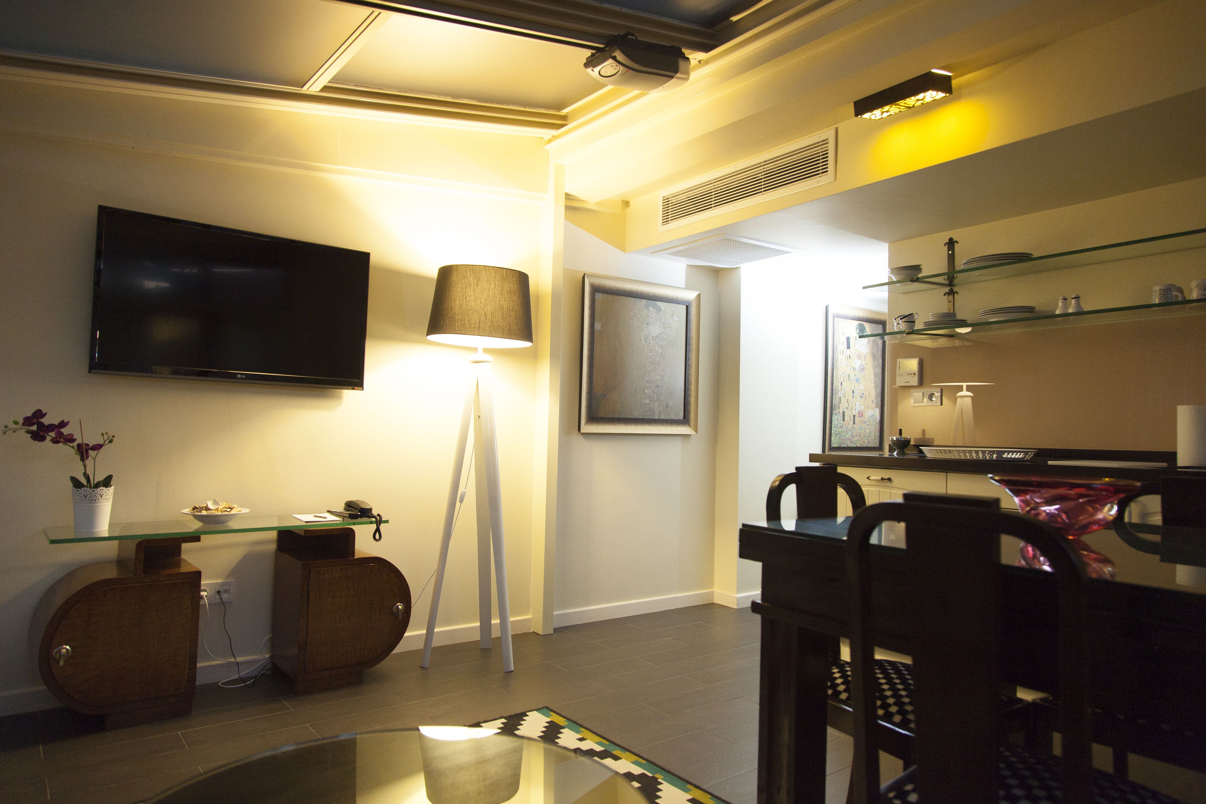 A modern living room with a flat-screen TV, a glass coffee table, a lamp, and a dining area with chairs and a table.