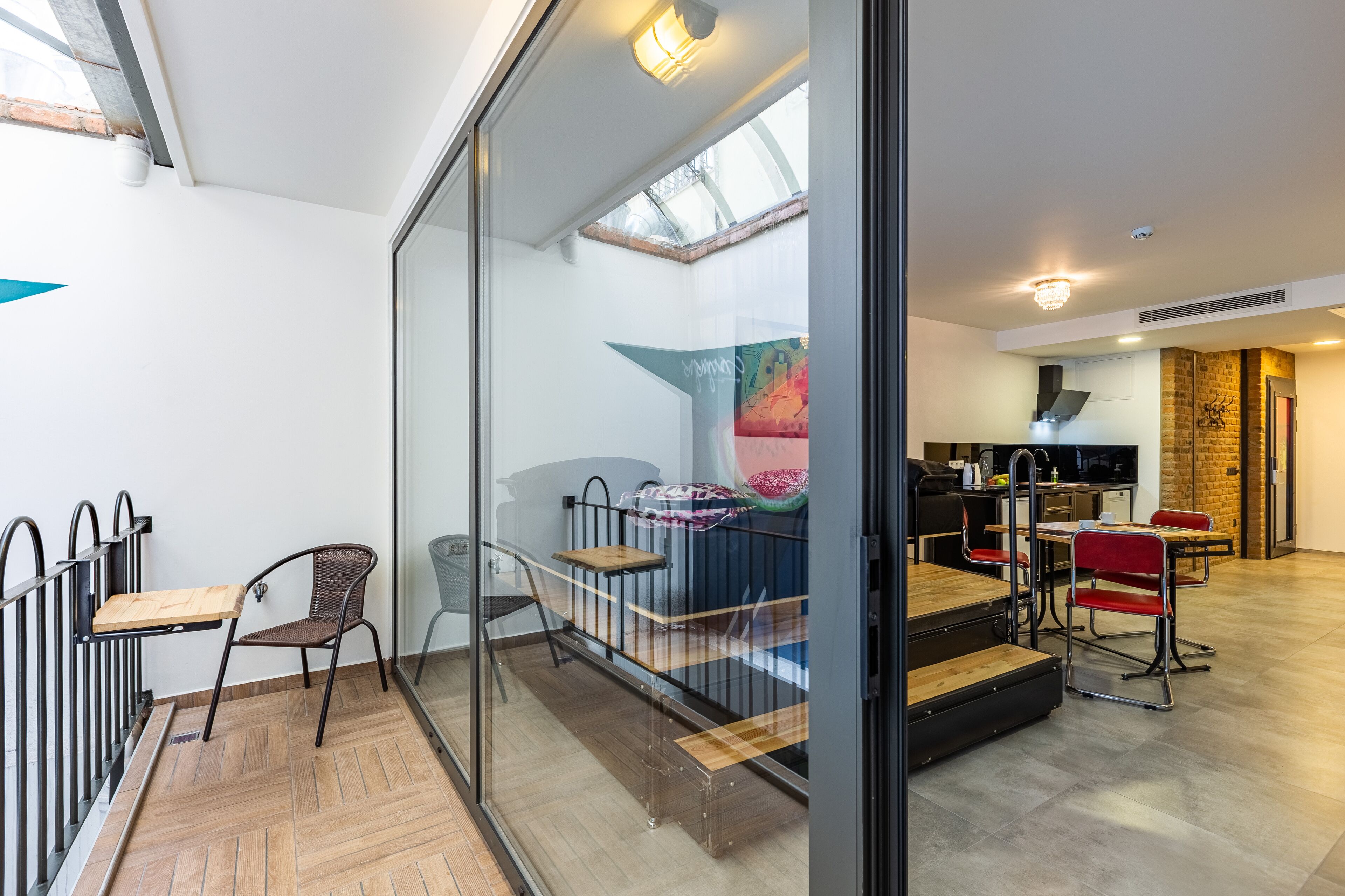 A modern interior with a glass partition, a dining area with red chairs, a kitchen with a brick wall, and a staircase with a wooden railing.