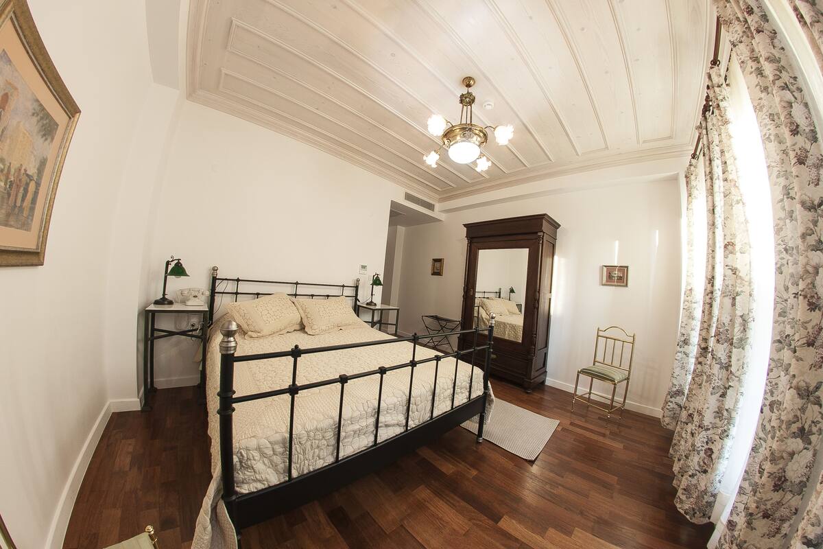 Ein Schlafzimmer mit einem großen Bett, einem Holzsessel, einem Nachttisch, einem Spiegel und einem Deckenventilator.
