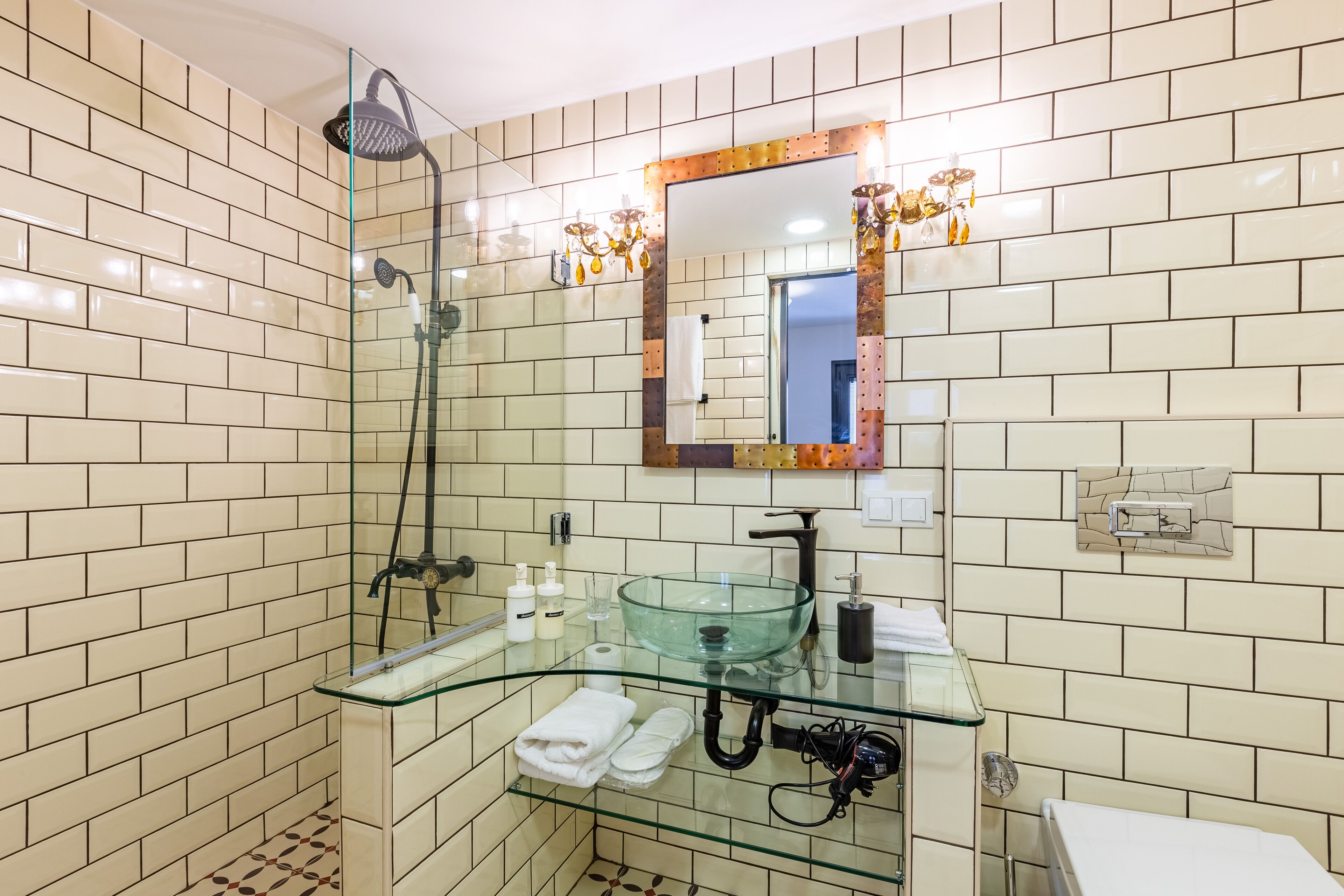 A bathroom with beige tiled walls, a glass shower enclosure, a glass sink with a round basin, and a mirror with a decorative frame.