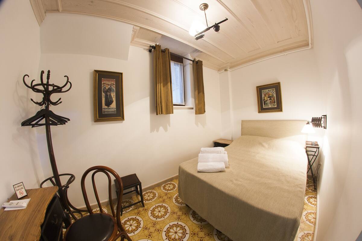 Ein Hotelzimmer mit einem Bett, einem Sessel, einem Kleiderhaken, einem kleinen Tisch und einem Fenster mit Vorhängen.