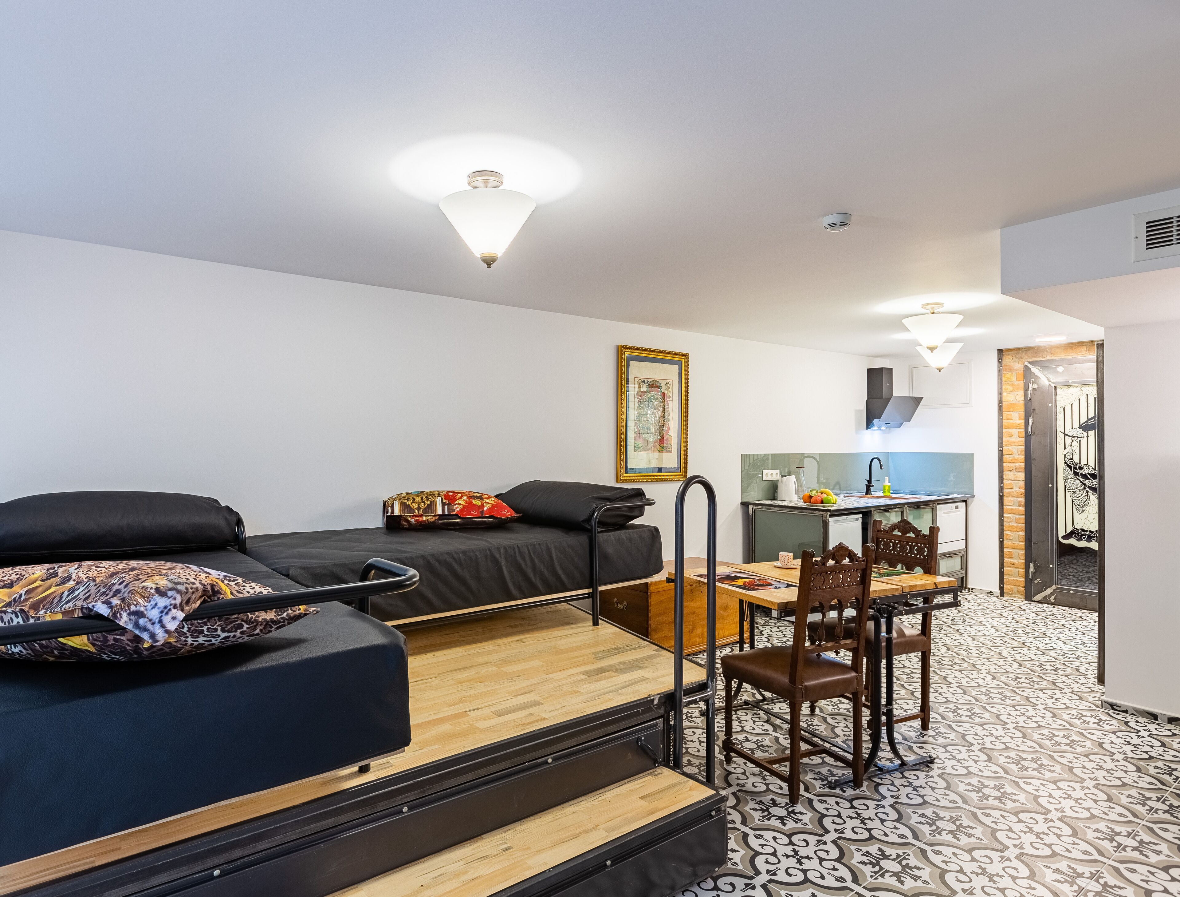 Quarto compacto com uma cama, uma escrivaninha e uma cadeira. Possui uma cozinha compacta com pia e fogão. O quarto tem um piso estampado e uma luminária no teto.