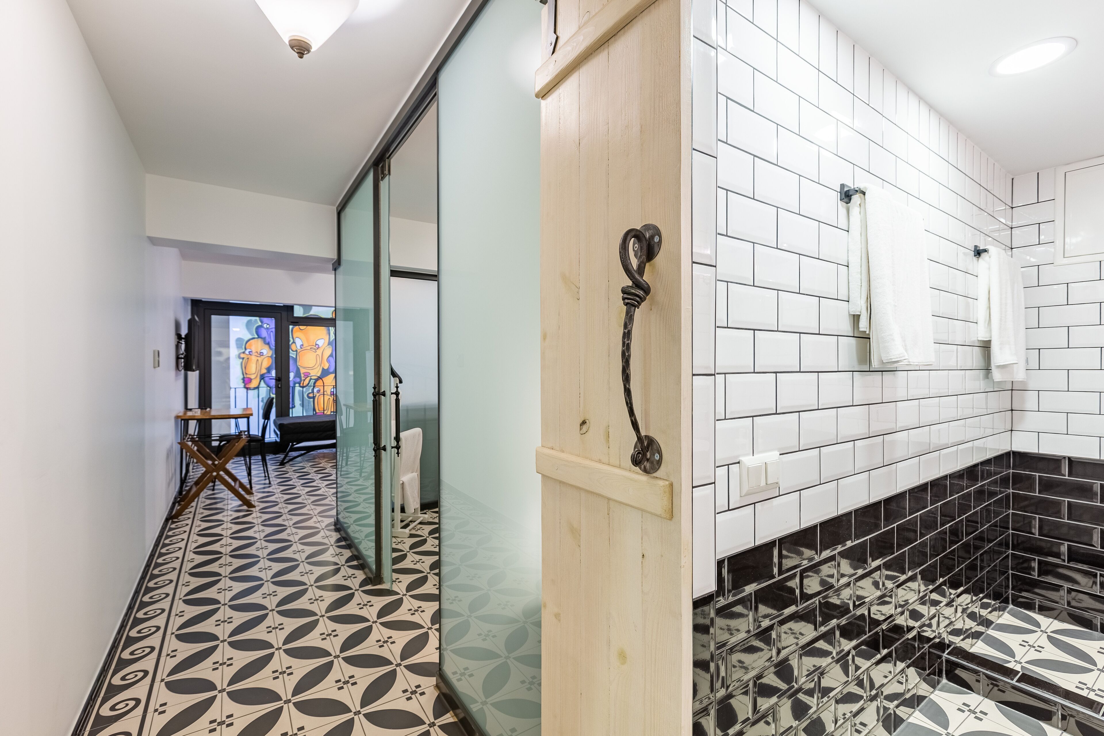 A modern bathroom with white tiles, a black and white patterned floor, and a wooden door with a metal handle.