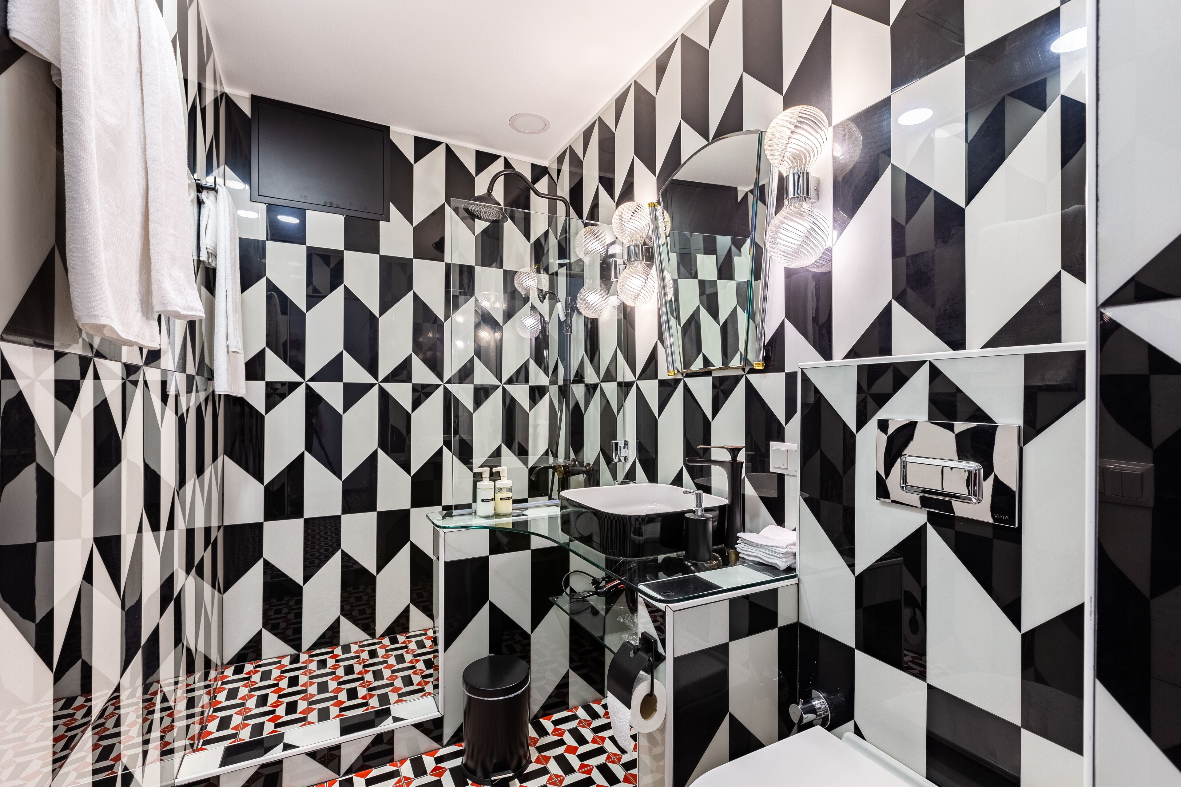 A modern bathroom with black and white geometric tiles, a glass sink, and a shower with multiple heads.