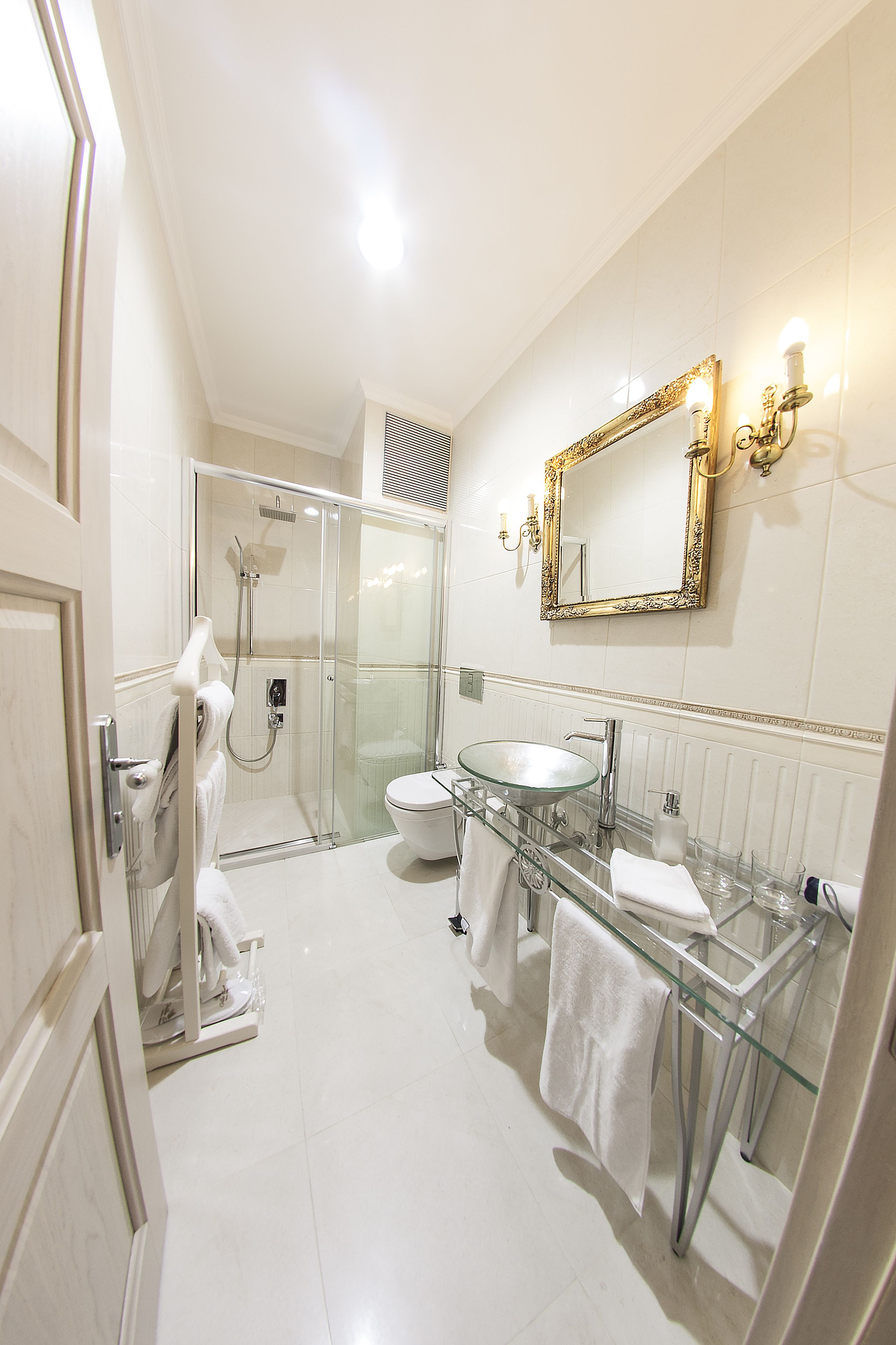 A modern bathroom with a glass shower enclosure, a white toilet, a glass sink, and a large mirror with a gold frame.