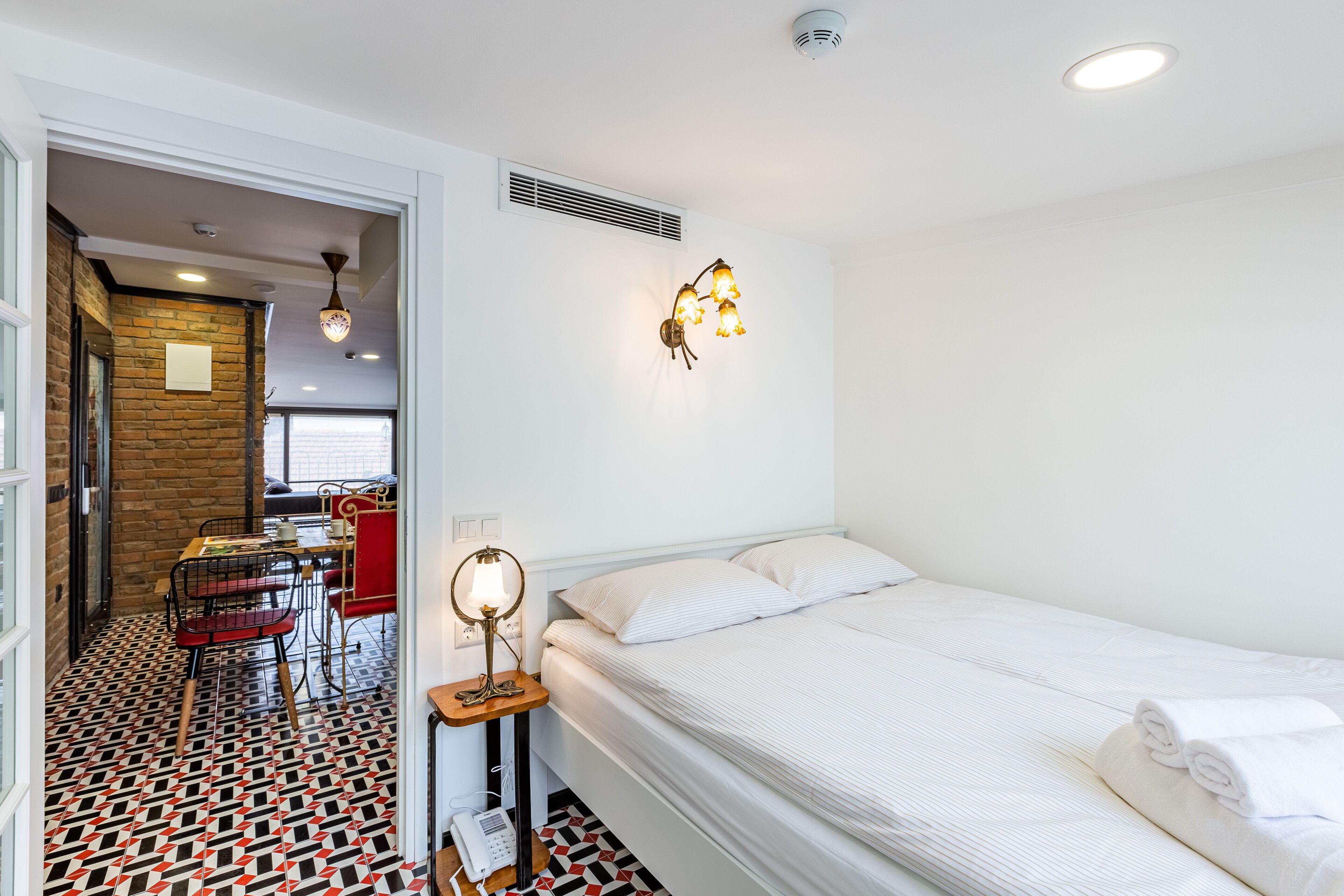A hotel room with a bed, a desk with a lamp, a chair, a patterned floor, and a view of the outside.