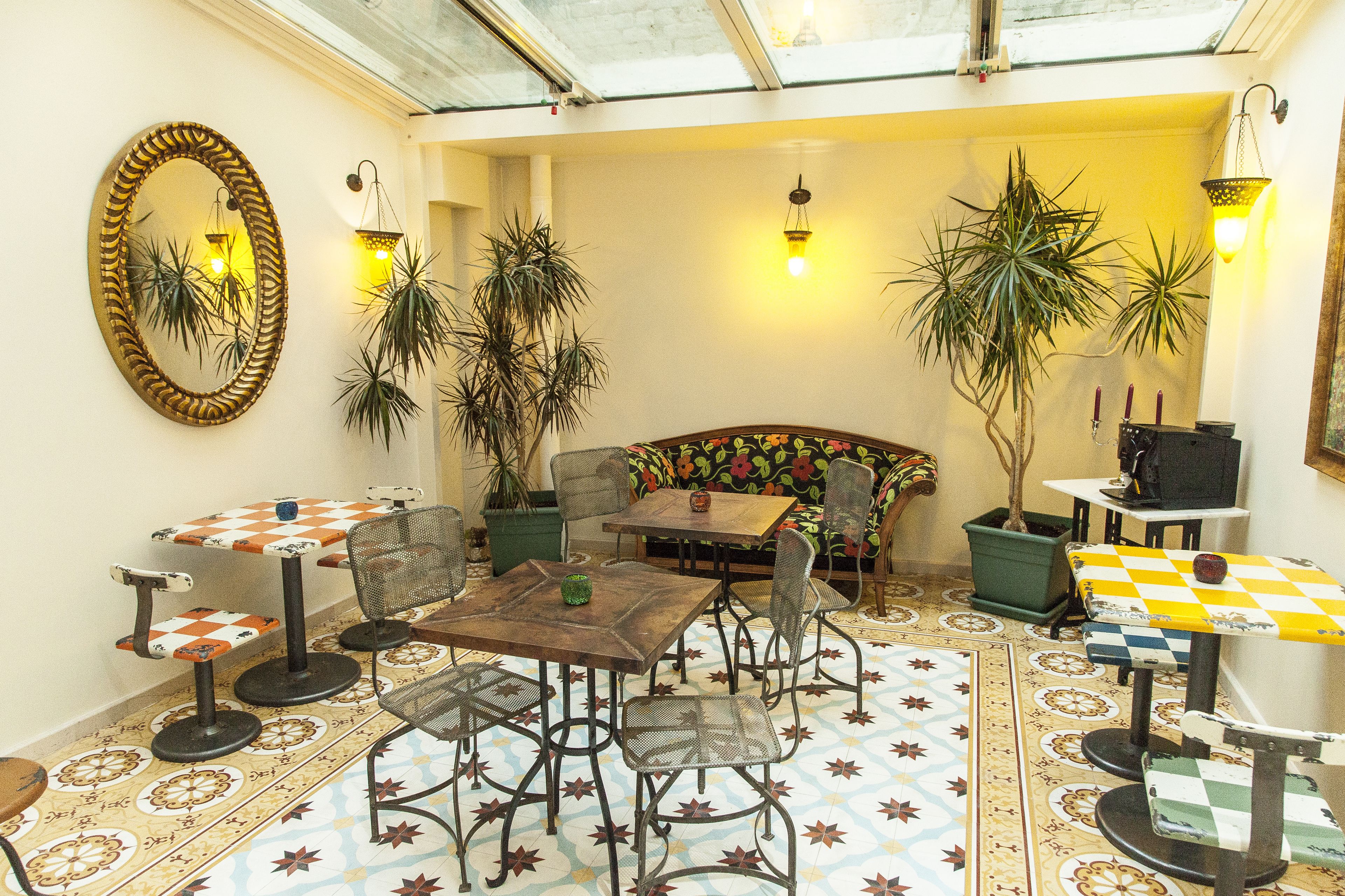 Un área de estar interior acogedor con una mesa de madera, sillas de metal y un sofá. Hay plantas en macetas y un espejo en las paredes.